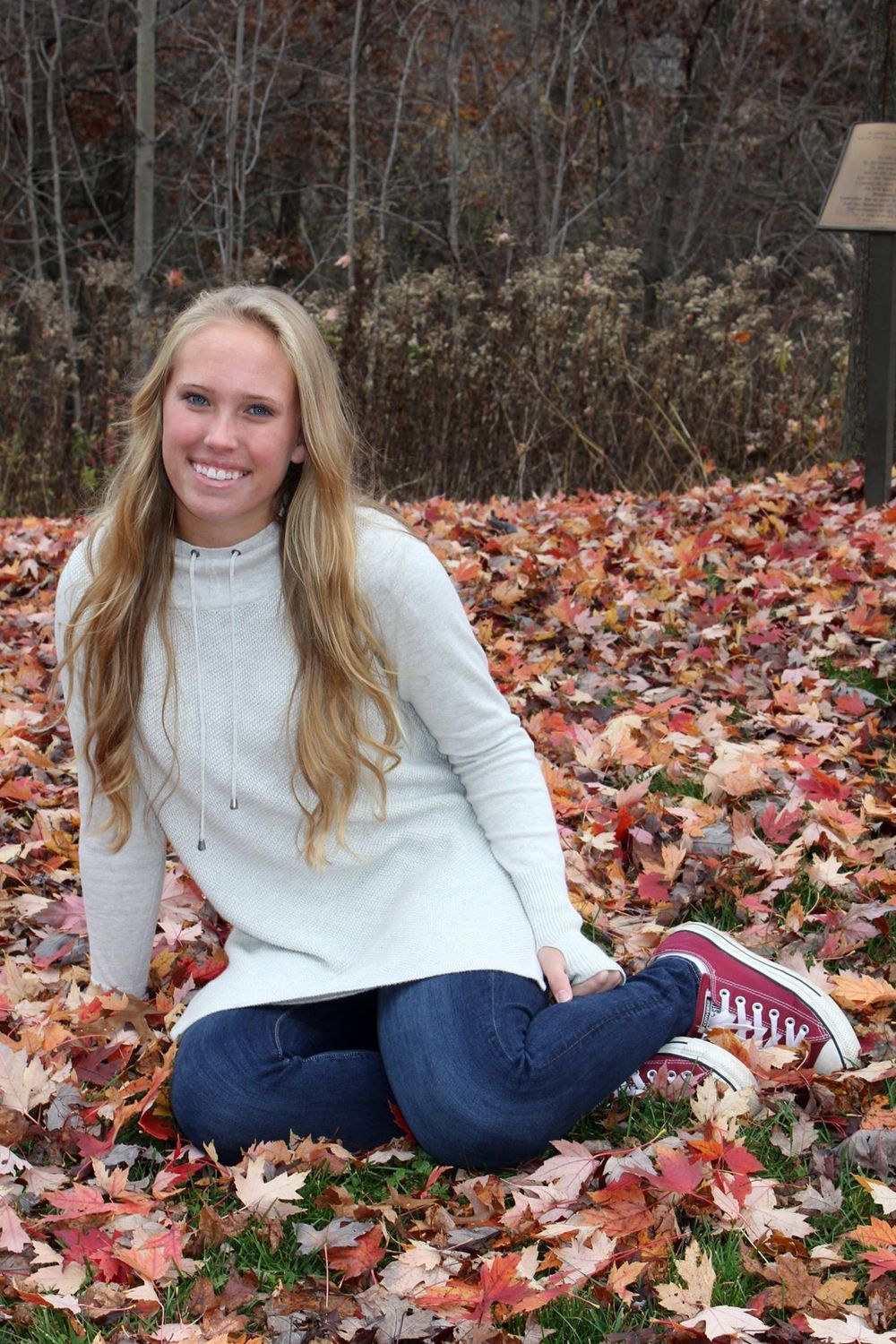 Smiling blonde teen girl sitting on fall leaves wearing white hoodie, jeans, and red Converse sneakers outdoors.