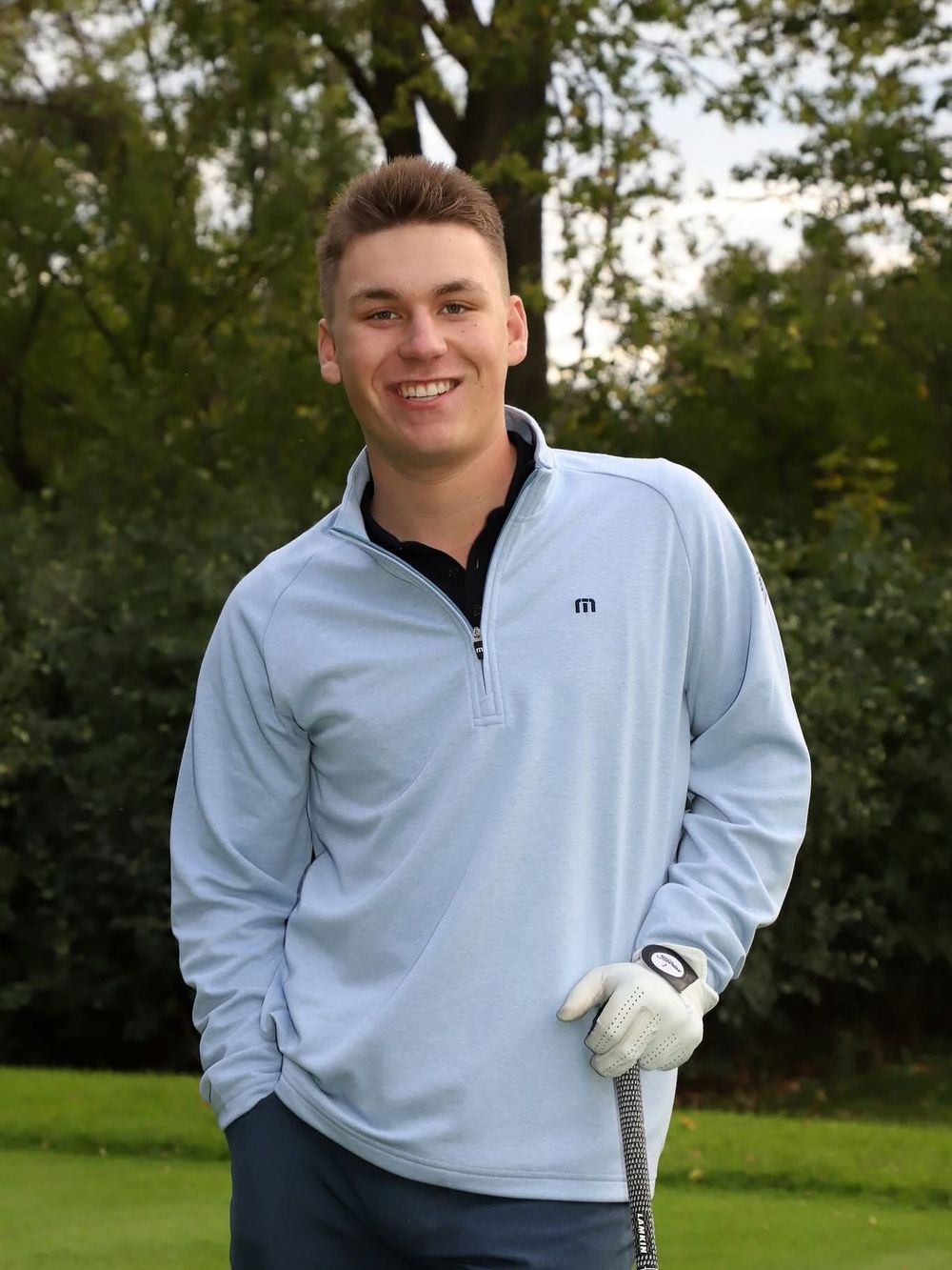 Young male golfer smiling outdoors, wearing a light blue Travis Mathew quarter-zip pullover and golf glove, holding a club.