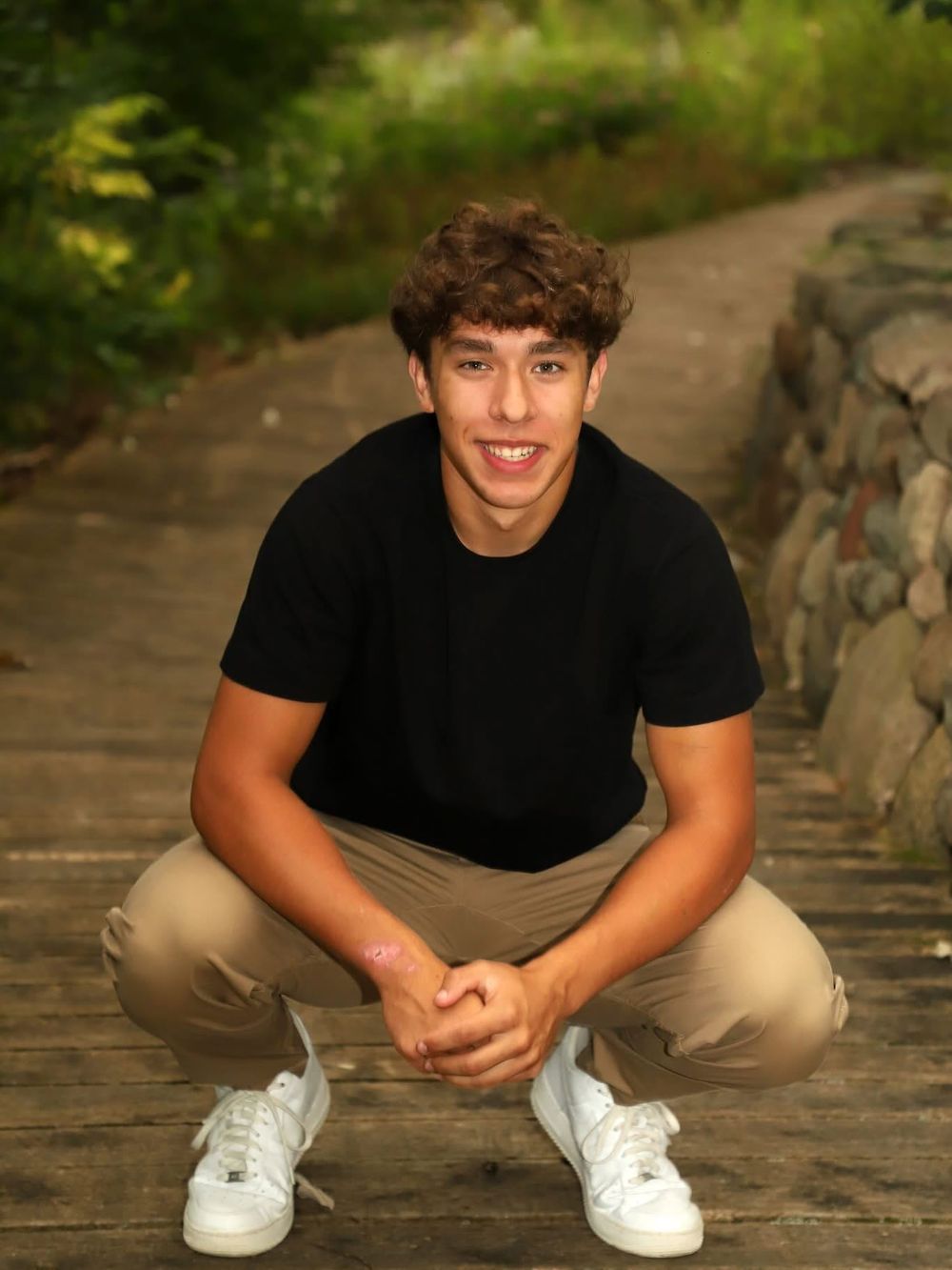 Young man in black t-shirt and khaki pants crouching on a wooden path surrounded by lush greenery.