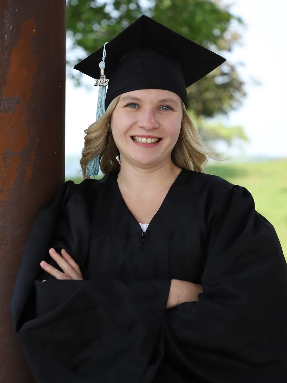 Smiling graduate in black cap and gown poses with arms crossed outdoors on a sunny day.