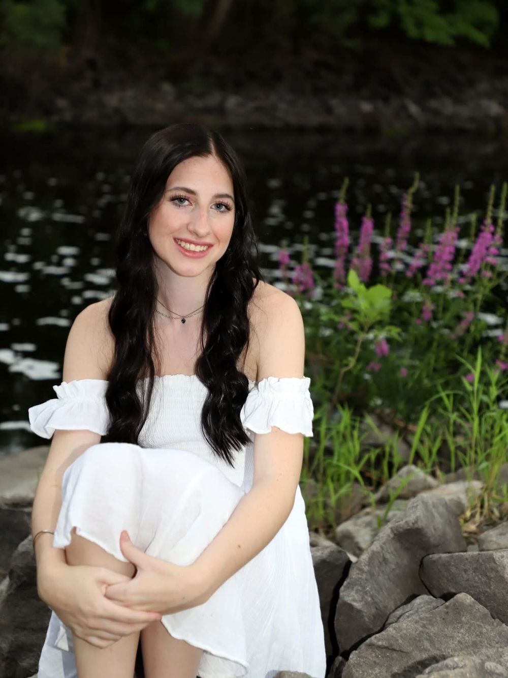 Young woman in white off-shoulder dress sitting on rocks by a pond with purple wildflowers in background.