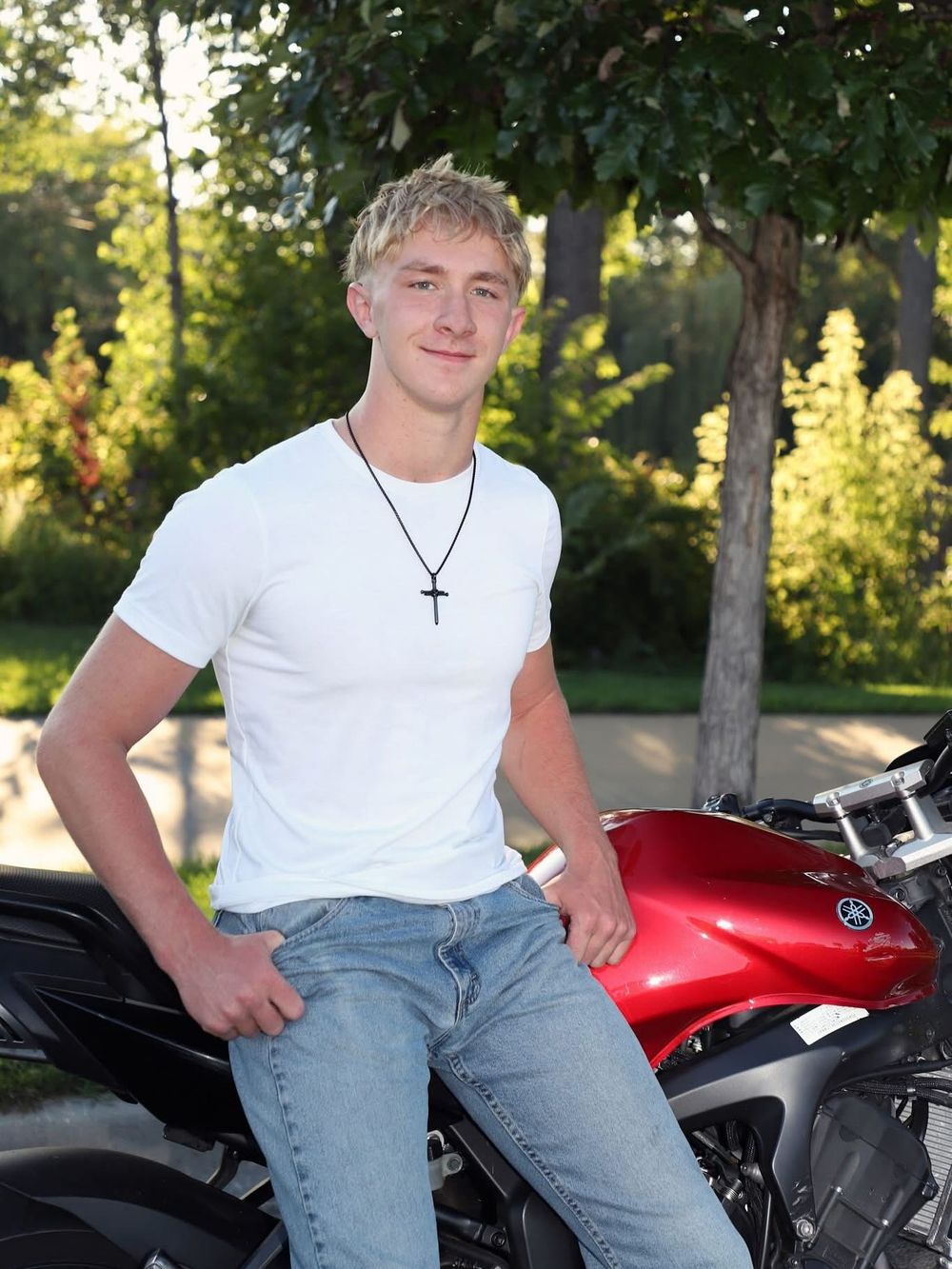 Young blond man in white t-shirt and jeans wearing cross necklace leaning against red Yamaha motorcycle outdoors.
