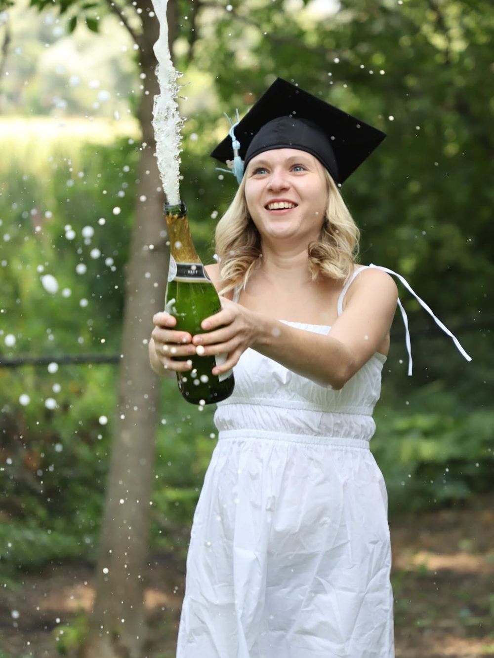 Smiling female graduate in white dress and cap celebrating by popping champagne bottle outdoors.