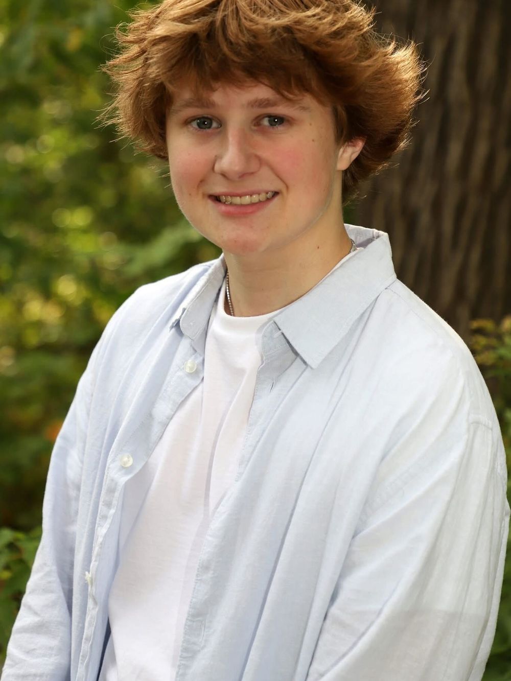 Smiling young person with red hair wearing a light blue shirt outdoors with green trees in the background.
