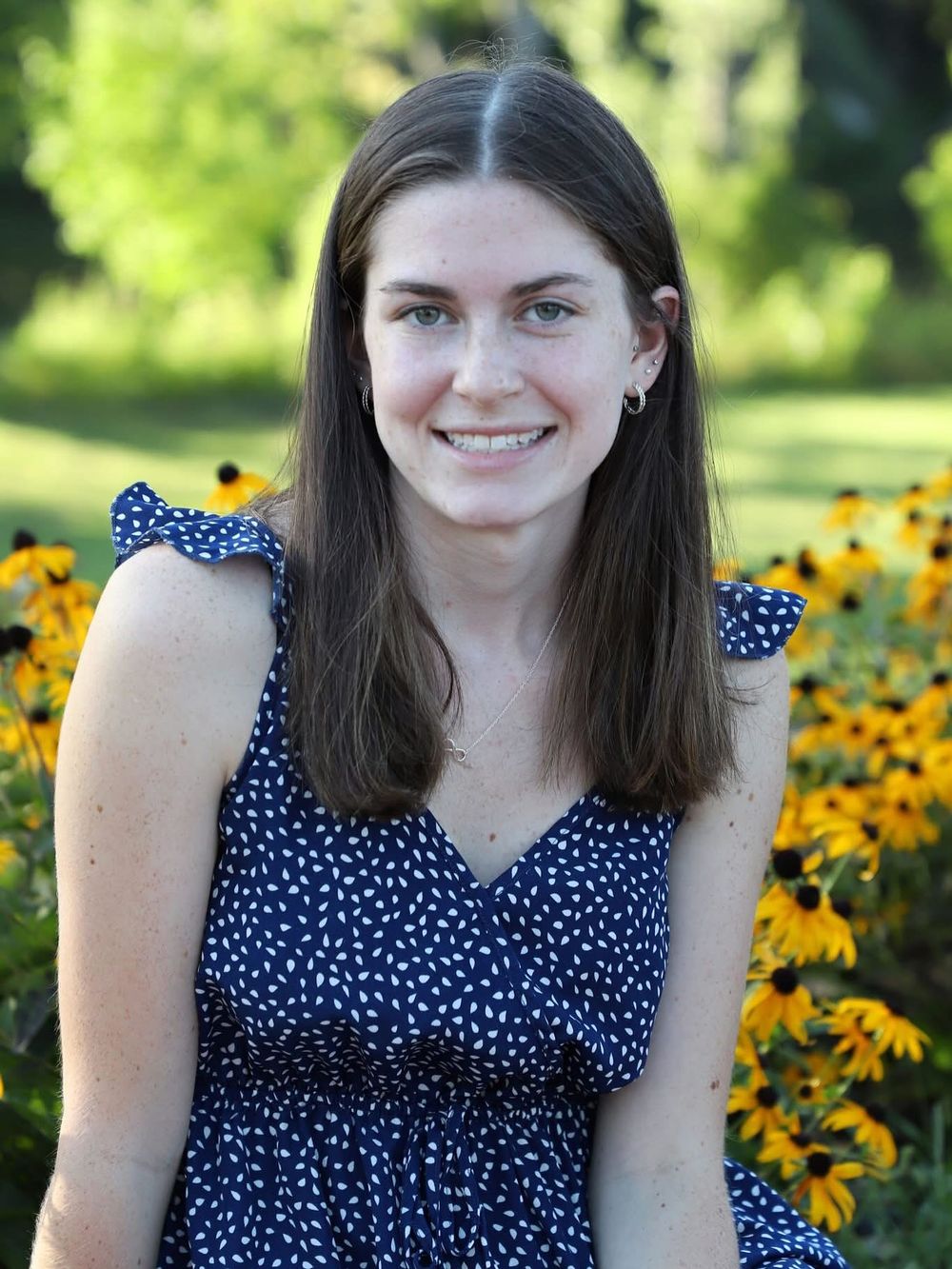 Young woman smiling in navy polka dot dress, posing outdoors among bright yellow black-eyed Susan flowers.
