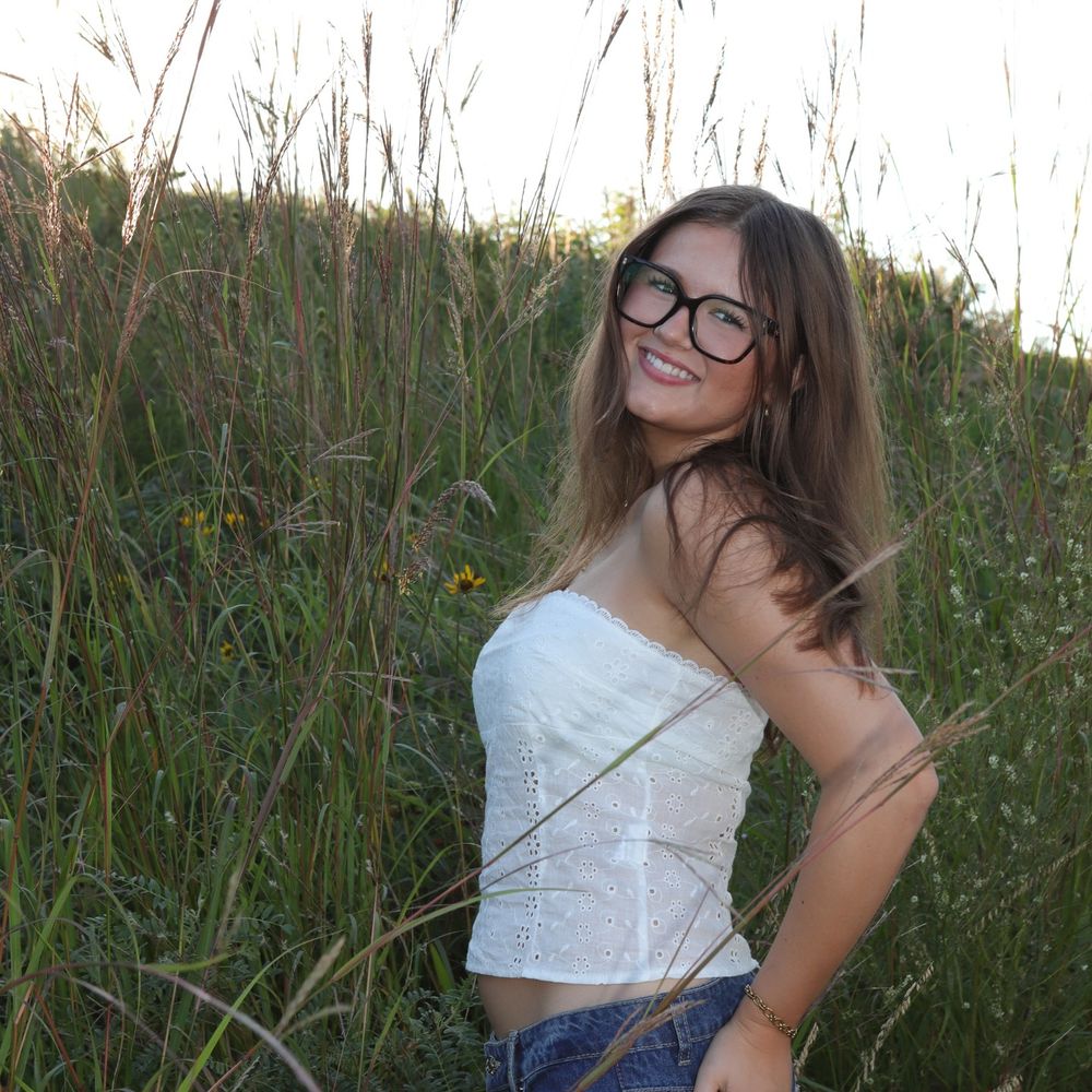 Young woman with glasses smiling in a white crop top and jeans, standing in a lush green field on a sunny day.