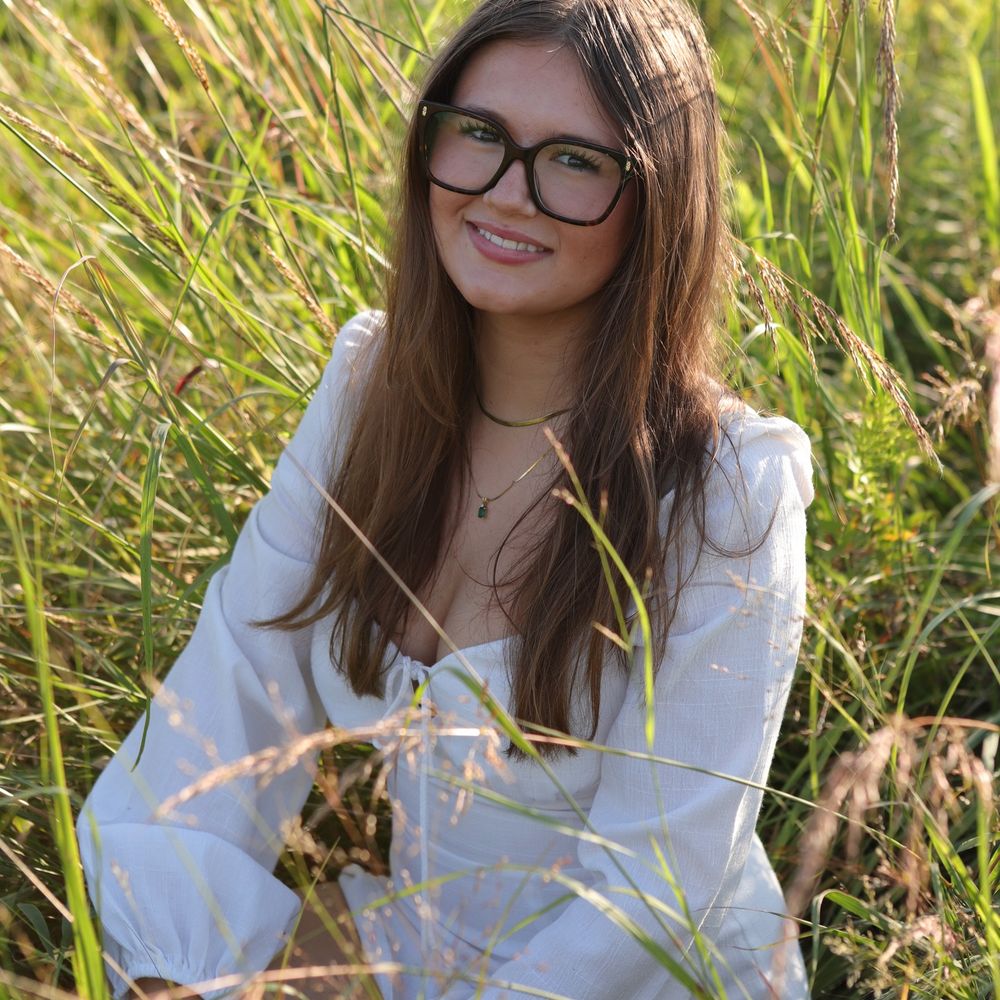 Young woman with glasses sitting in a sunlit grassy field, wearing a white blouse, smiling warmly.