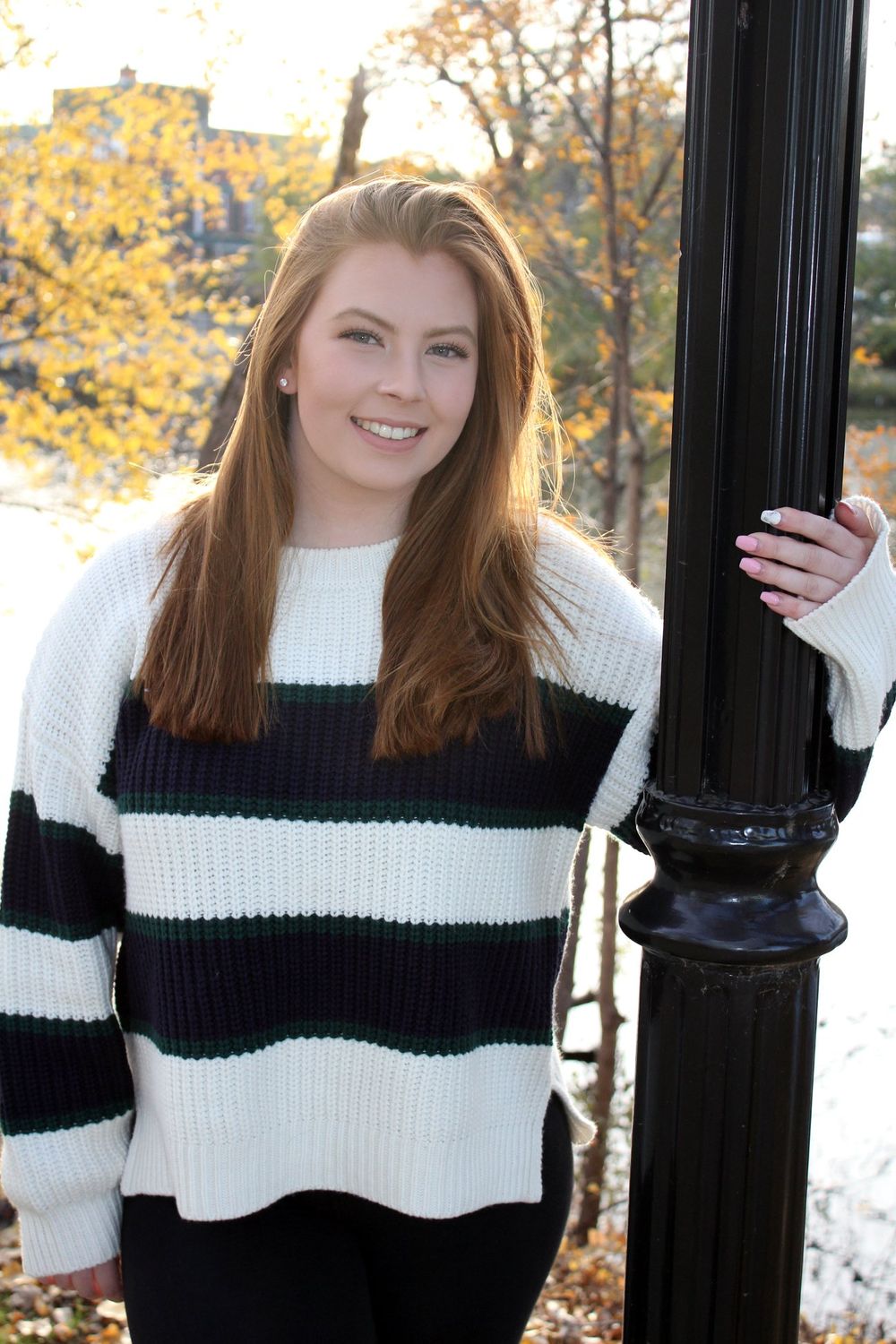 Young woman in striped sweater smiling beside black lamppost with autumn foliage background.