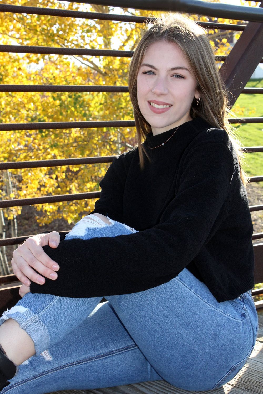 Young woman in black sweater and ripped jeans sitting on a bridge, smiling, with colorful autumn foliage behind her.