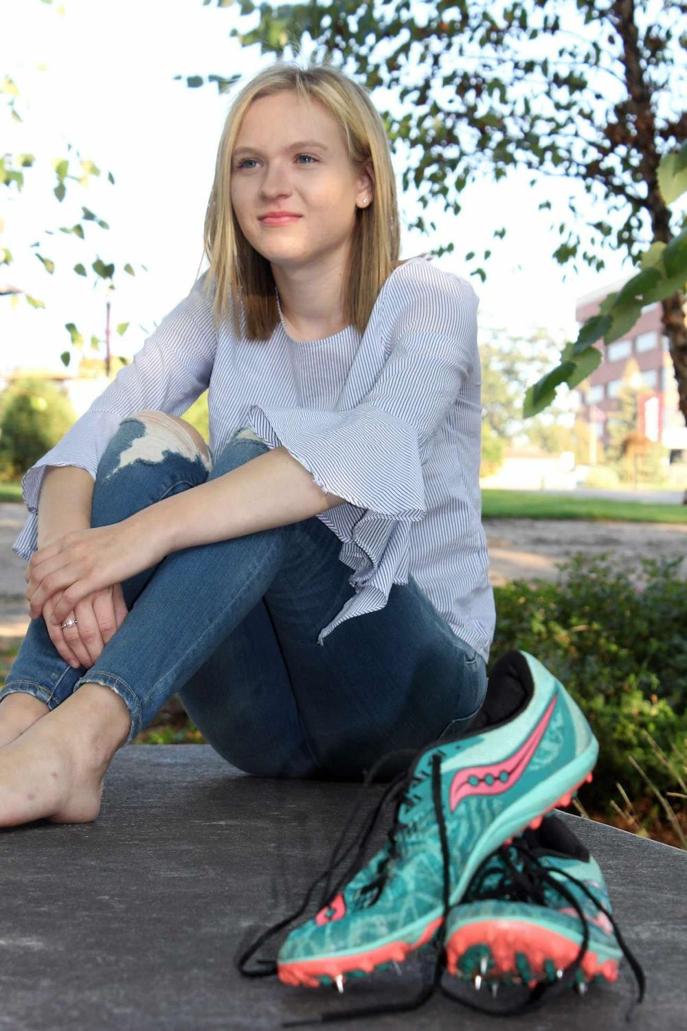 Young blonde woman sitting outdoors in ripped jeans and white top with teal running spikes beside her.