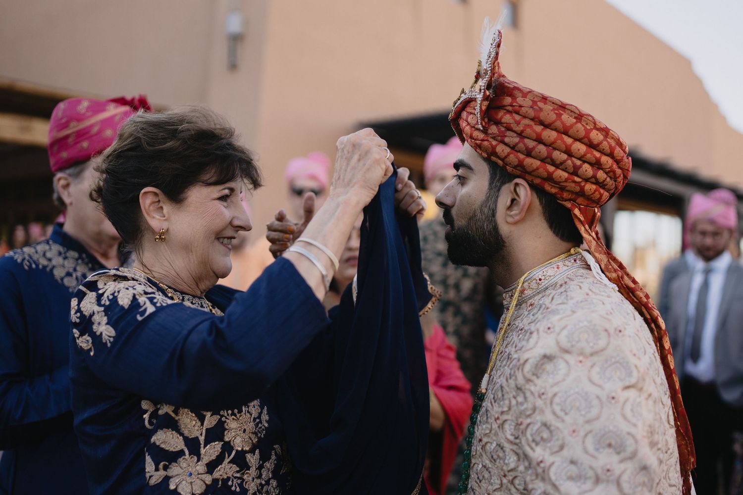 Elderly woman in blue dress adjusts groom's red turban during an intimate Indian wedding ceremony moment.at Sandia Resort and Casino