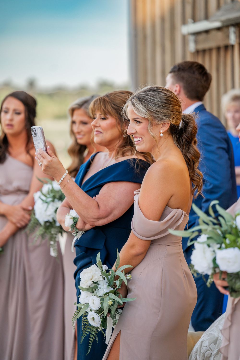 Bridesmaids in mauve dresses hold white bouquets while standing in line during an outdoor wedding ceremony.