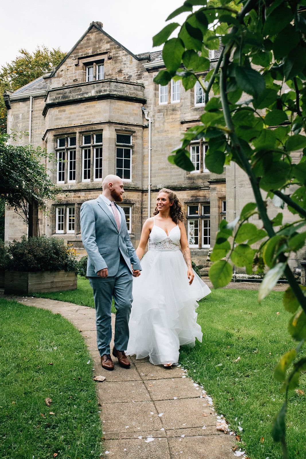 A couple in wedding attire dances joyfully along a stone path outside a historic manor house.