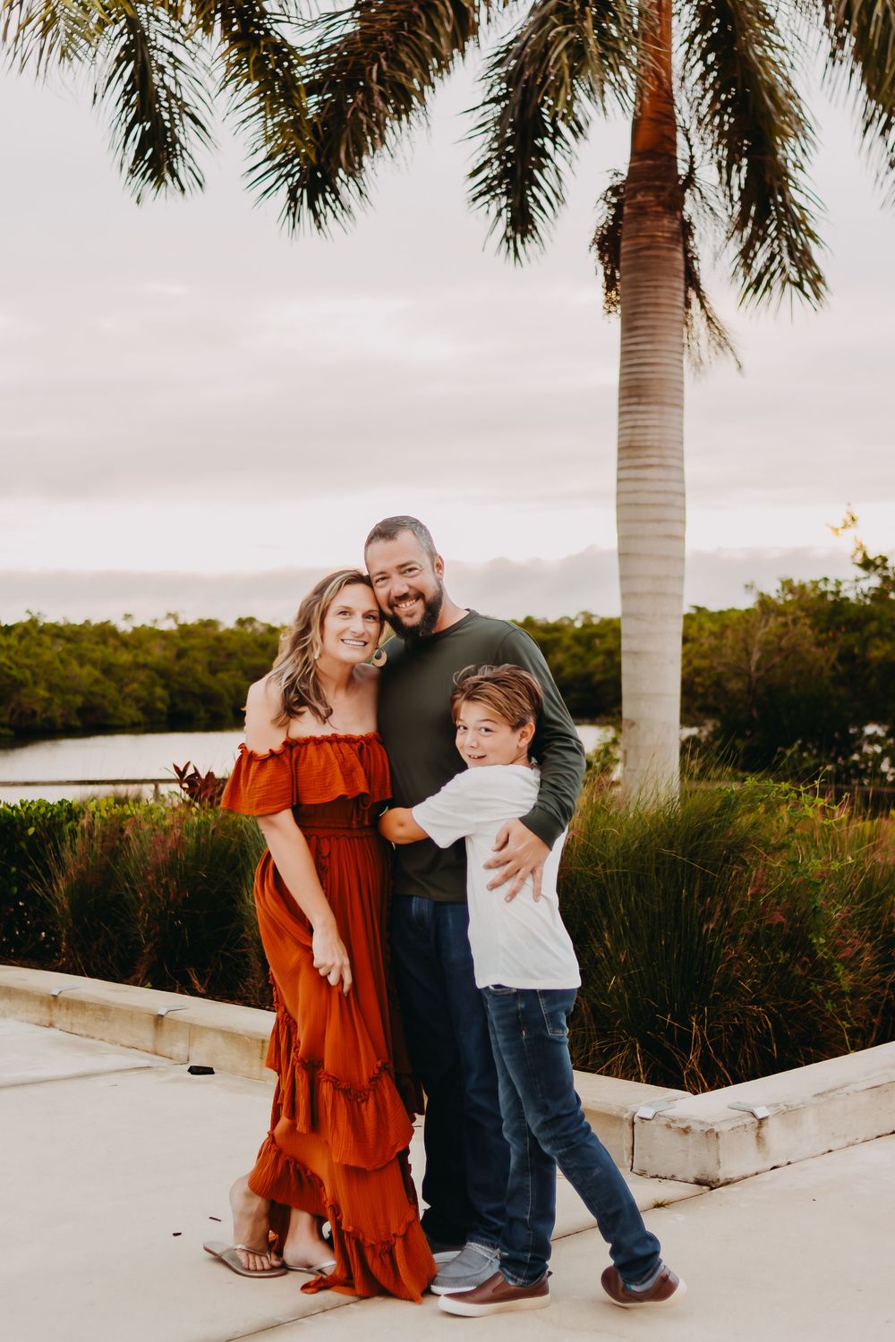 A family in coordinated outfits poses together on a white wall with palm trees and greenery in the background at sunset.
