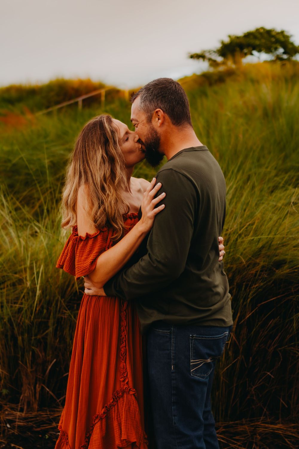 A couple embraces in tall grass during golden hour sunset, with the woman wearing a flowing red dress.