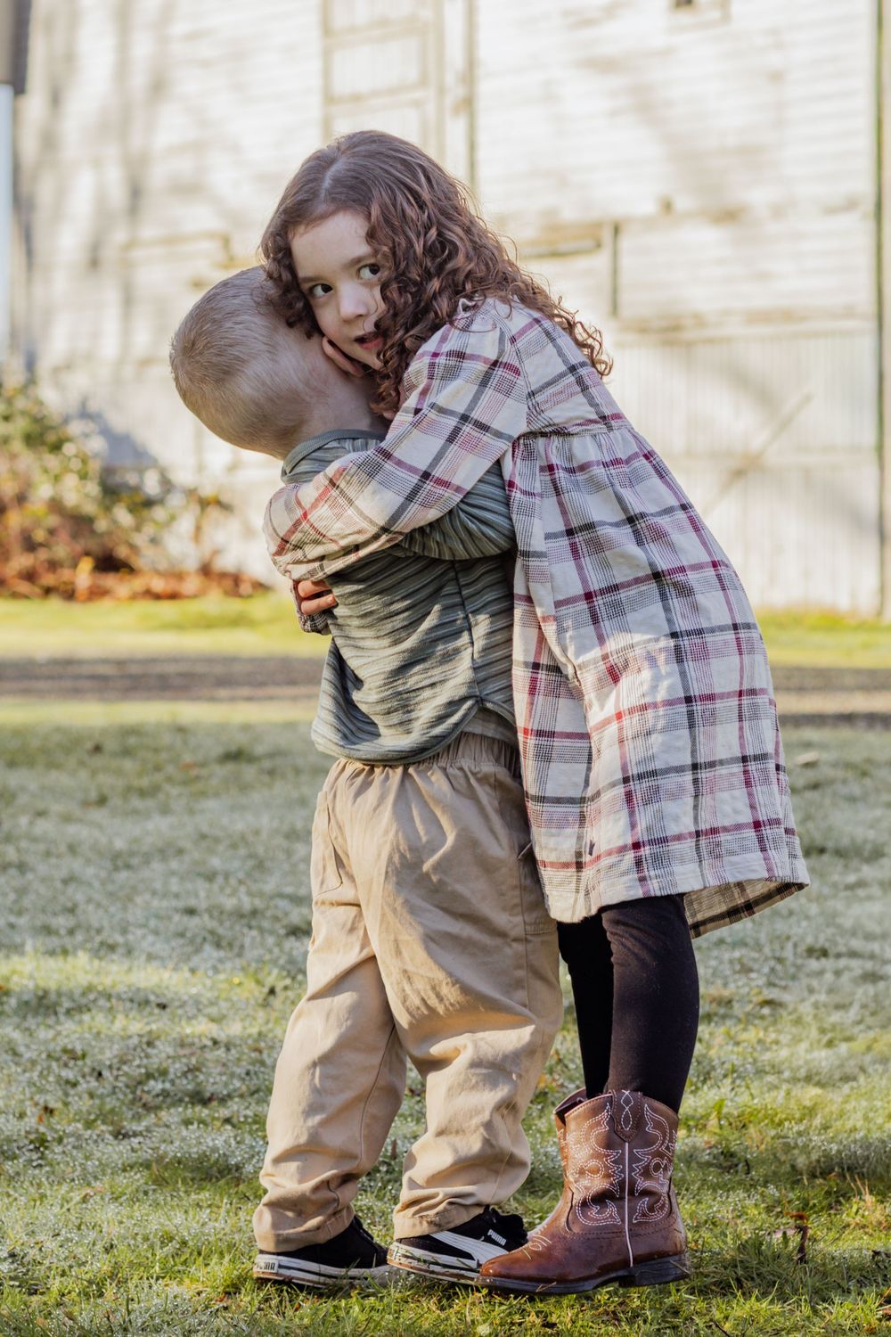 Young girl in plaid dress and cowboy boots hugs her little brother in a grassy outdoor setting.