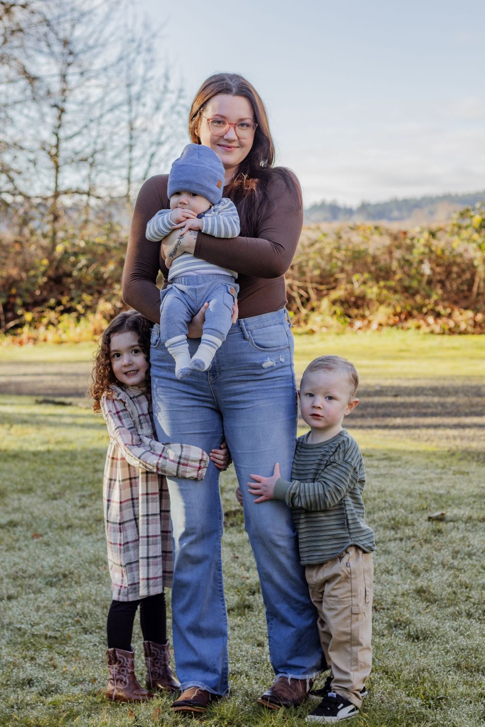 Smiling mother holds infant while two young children stand beside her in a sunny outdoor field.
