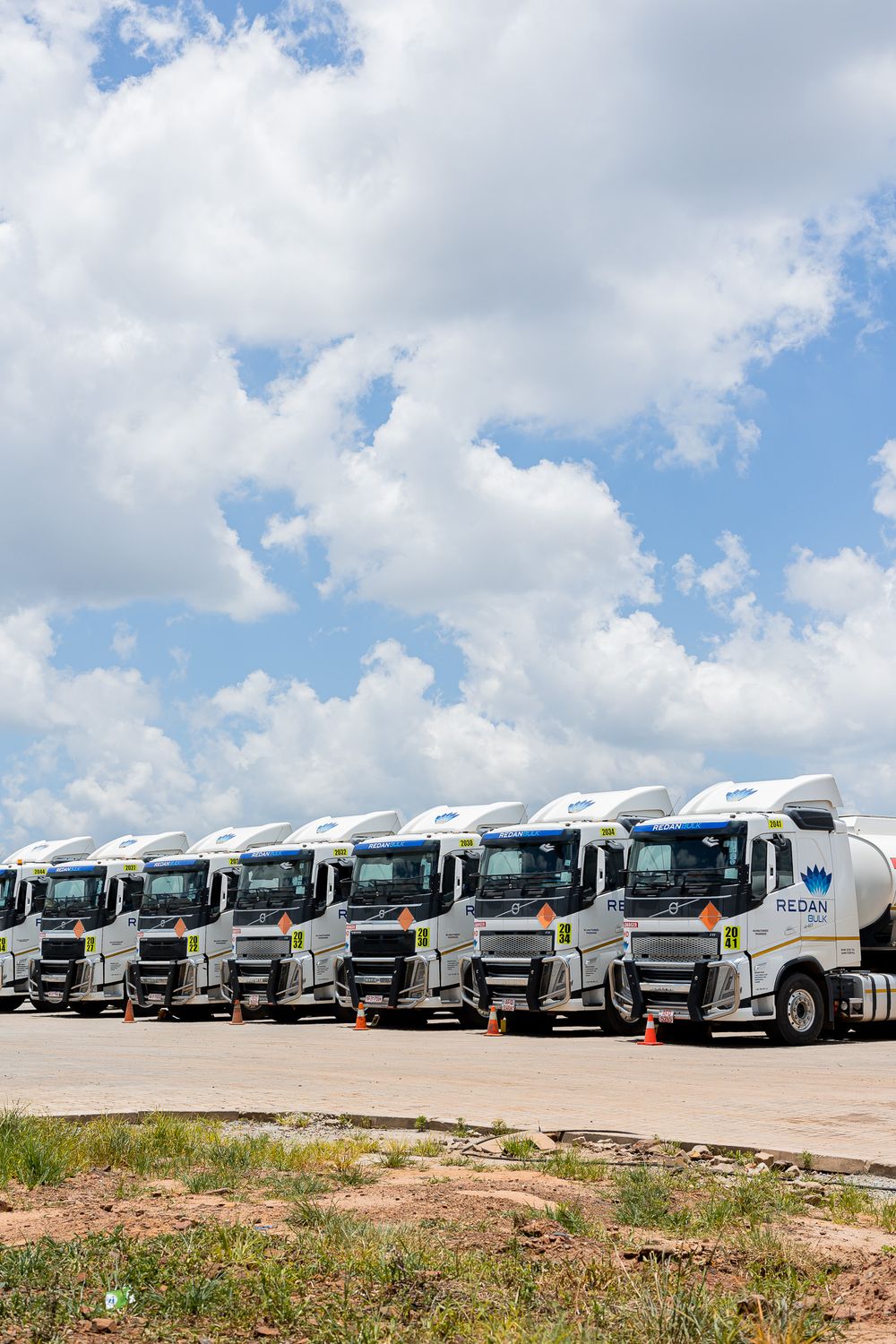 A row of white semi trucks and tankers parked in line on dirt ground against a cloudy blue sky.