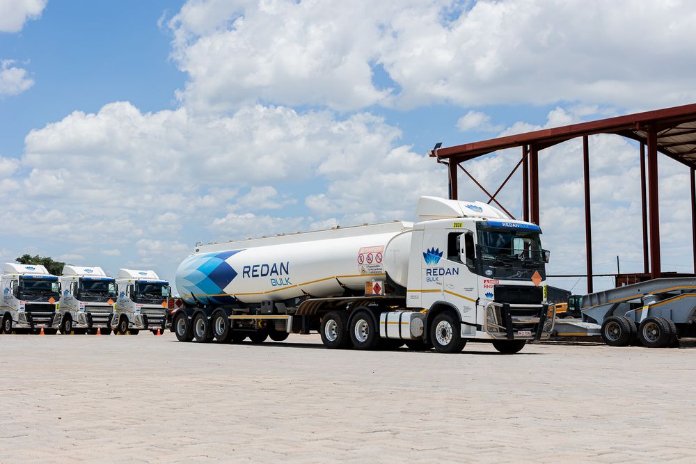 A row of white commercial tanker trucks parked in a line at a truck depot under a partly cloudy sky.