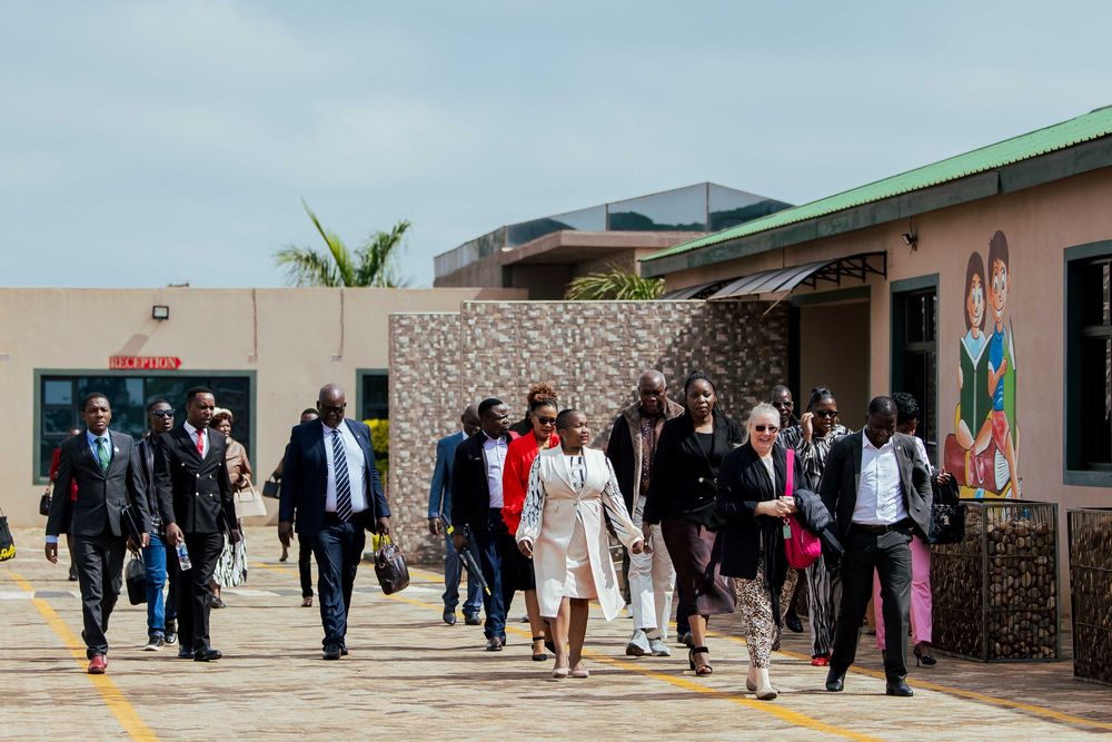 A line of people in formal attire walk along an outdoor corridor with palm trees and buildings in the background.