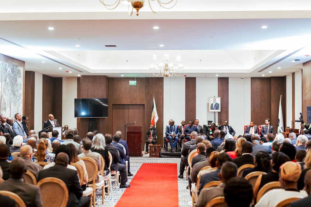 Conference attendees seated in rows face a stage with red carpet in an elegant hotel ballroom with wood paneled walls.