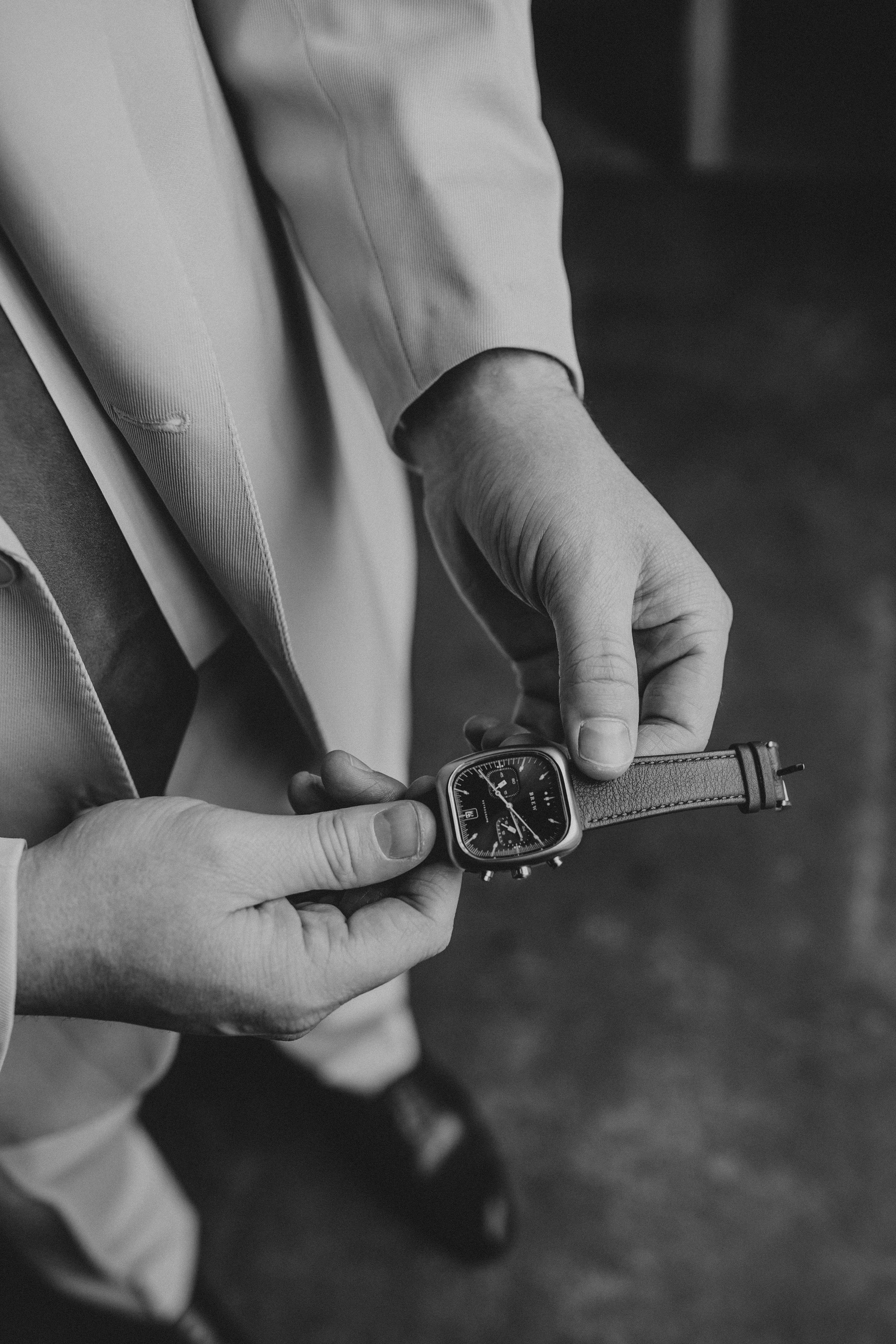 Black and white close-up of hands adjusting a leather watch strap against a formal suit background.