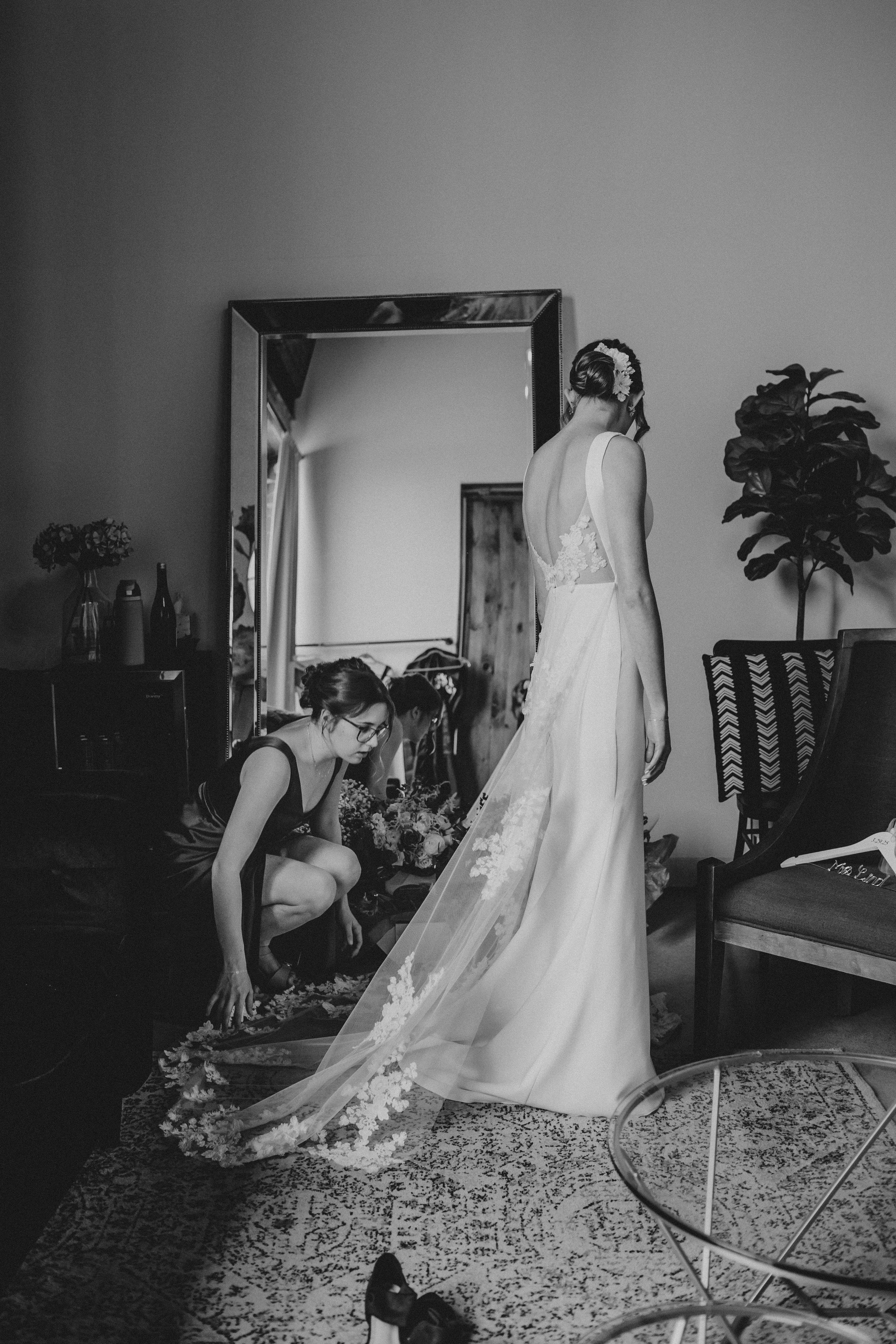 Black and white photo of a wedding dress being adjusted while someone stands in an elegant floor-length gown.