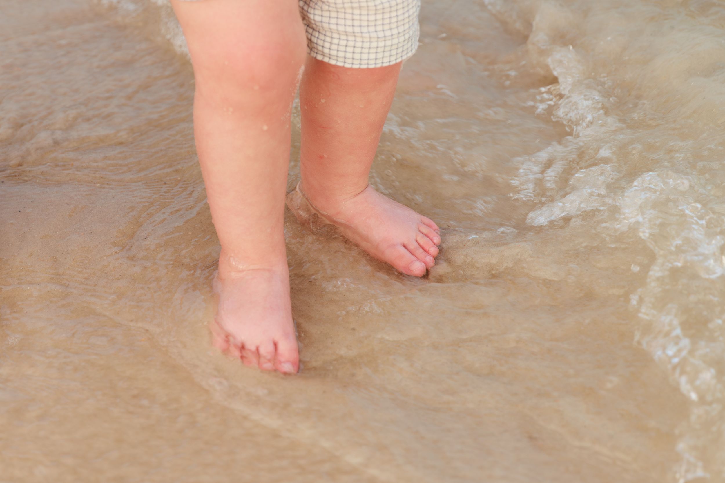 Tiny bare feet walk through shallow ocean water on a sandy beach as gentle waves roll in.