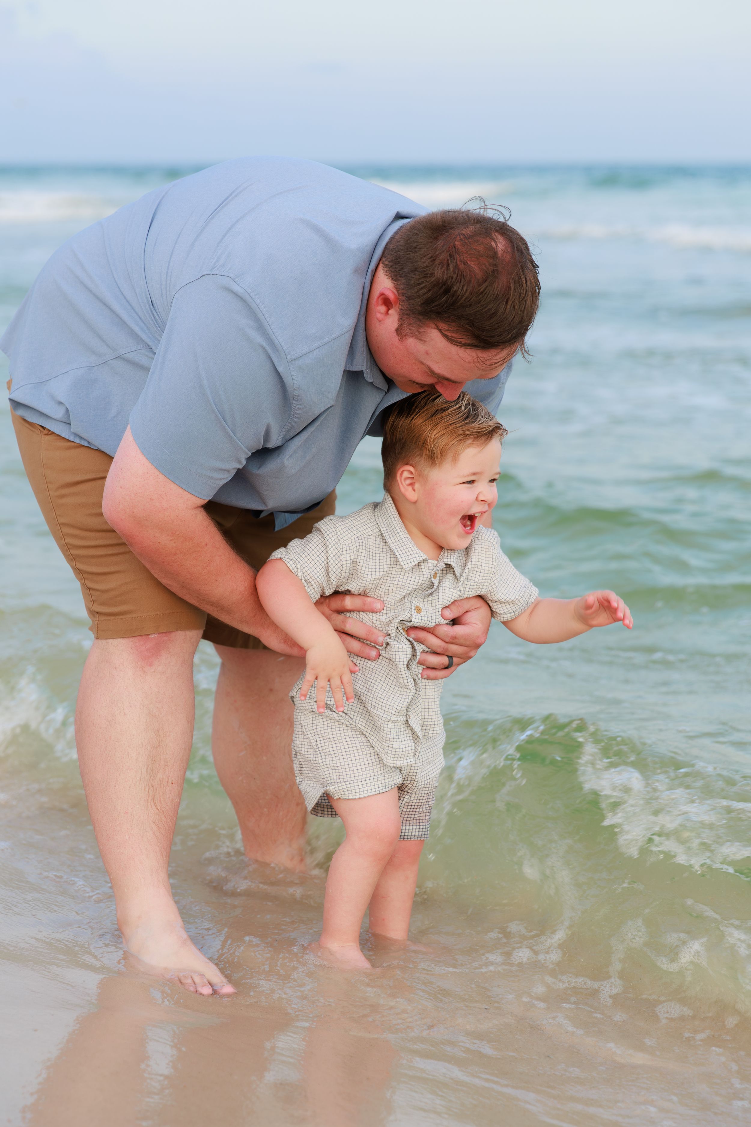 Parent and child playing together at the beach shoreline as gentle waves roll in on a sunny day.