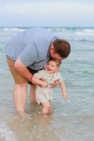 A parent and child play together in shallow ocean waves at the beach on a sunny day.