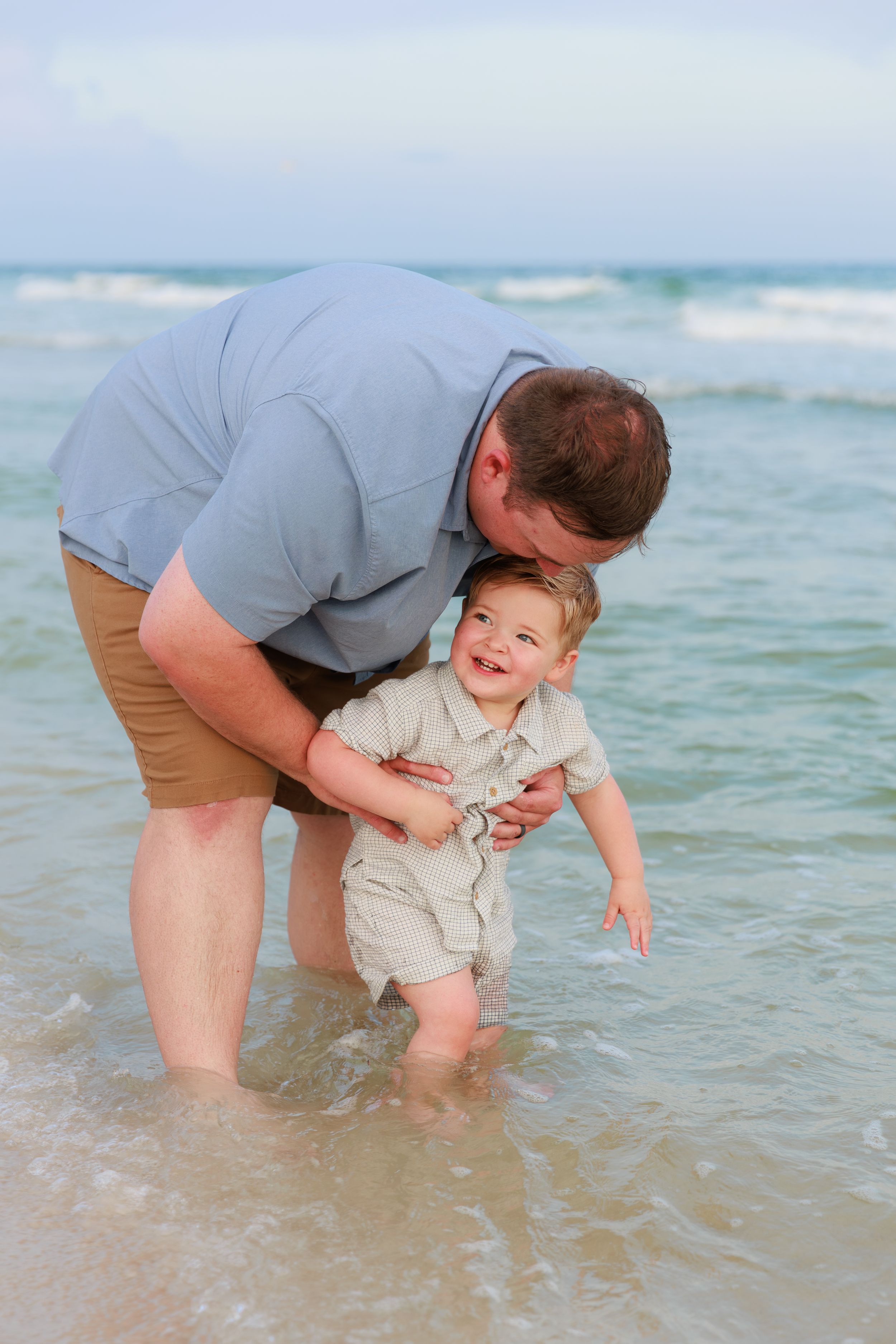 A parent helps a small child take their first steps in the gentle ocean waves at the beach.
