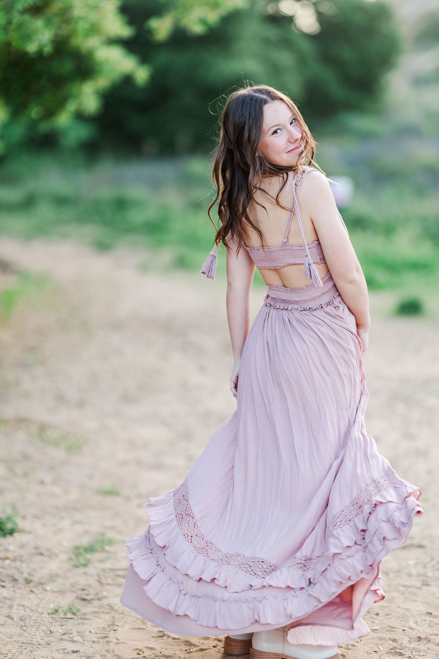 A person in a flowing lilac dress twirls playfully on a dirt path surrounded by lush green foliage.