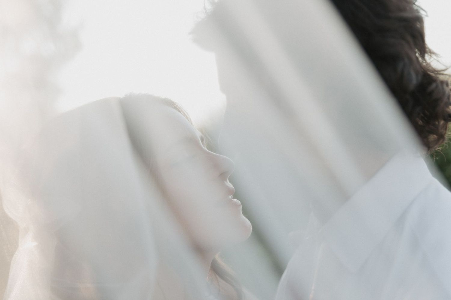 Dreamy soft-focus portrait of bride and groom nearly kissing beneath flowing white wedding veil.
