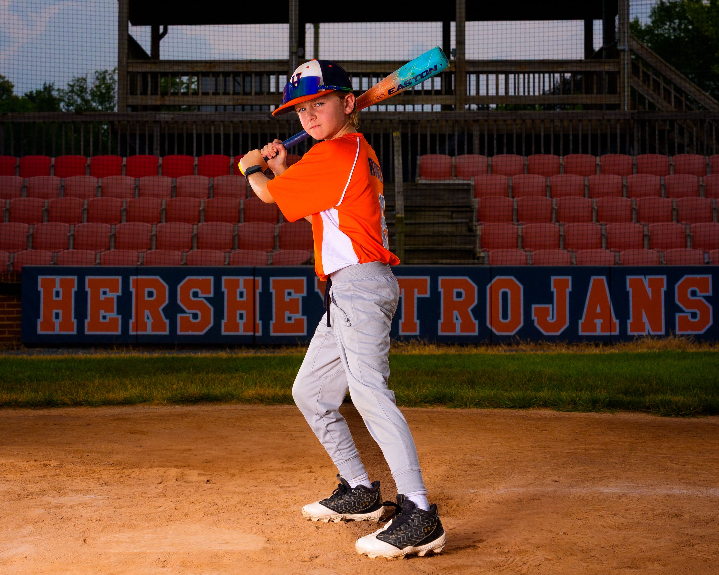 Youth baseball player in orange Hershey uniform, swinging bat on field, action portrait by SportsFox Photography.