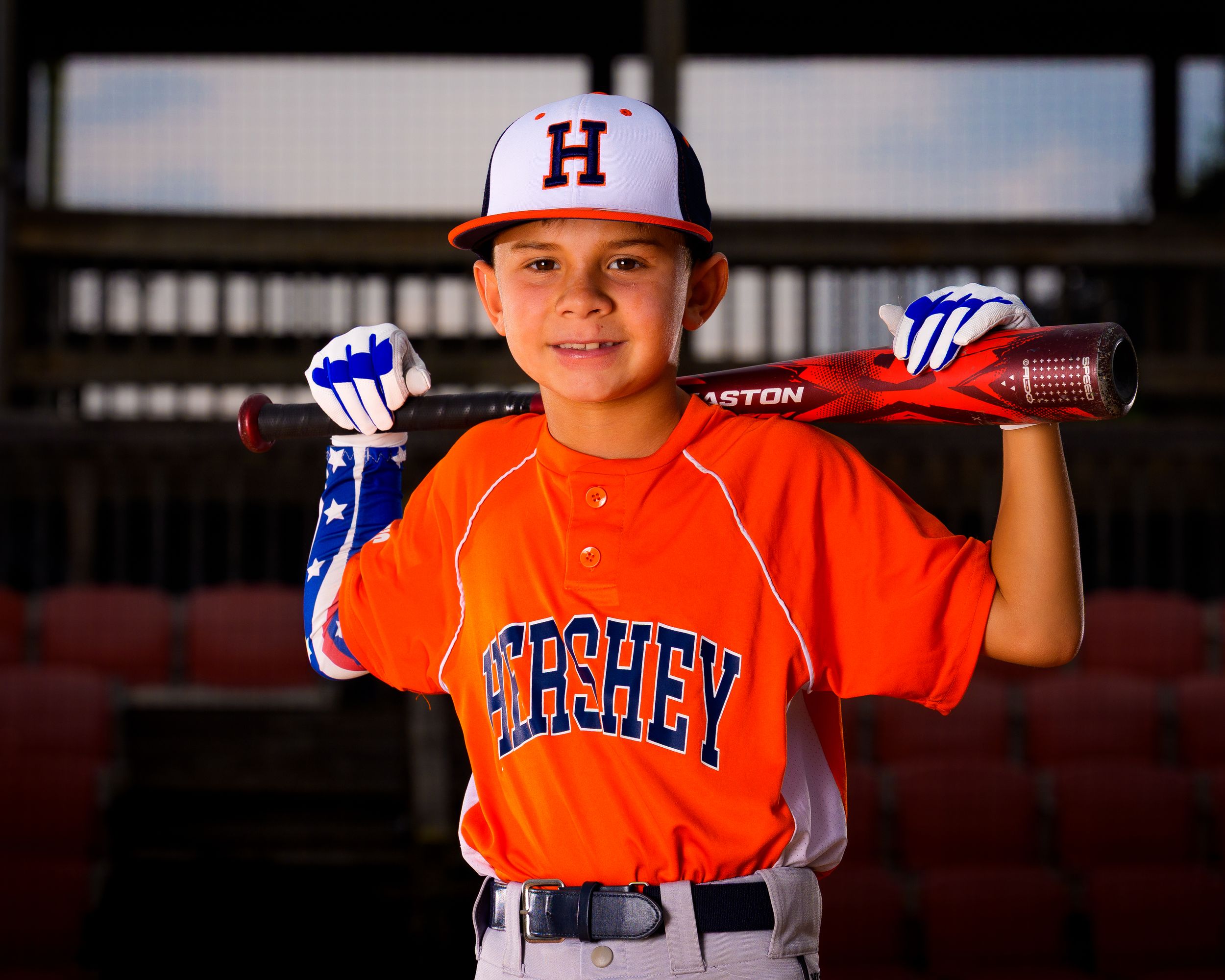 Little League baseball player in Hershey orange uniform, holding bat on shoulders and smiling, individual portrait by SportsFox Photography.