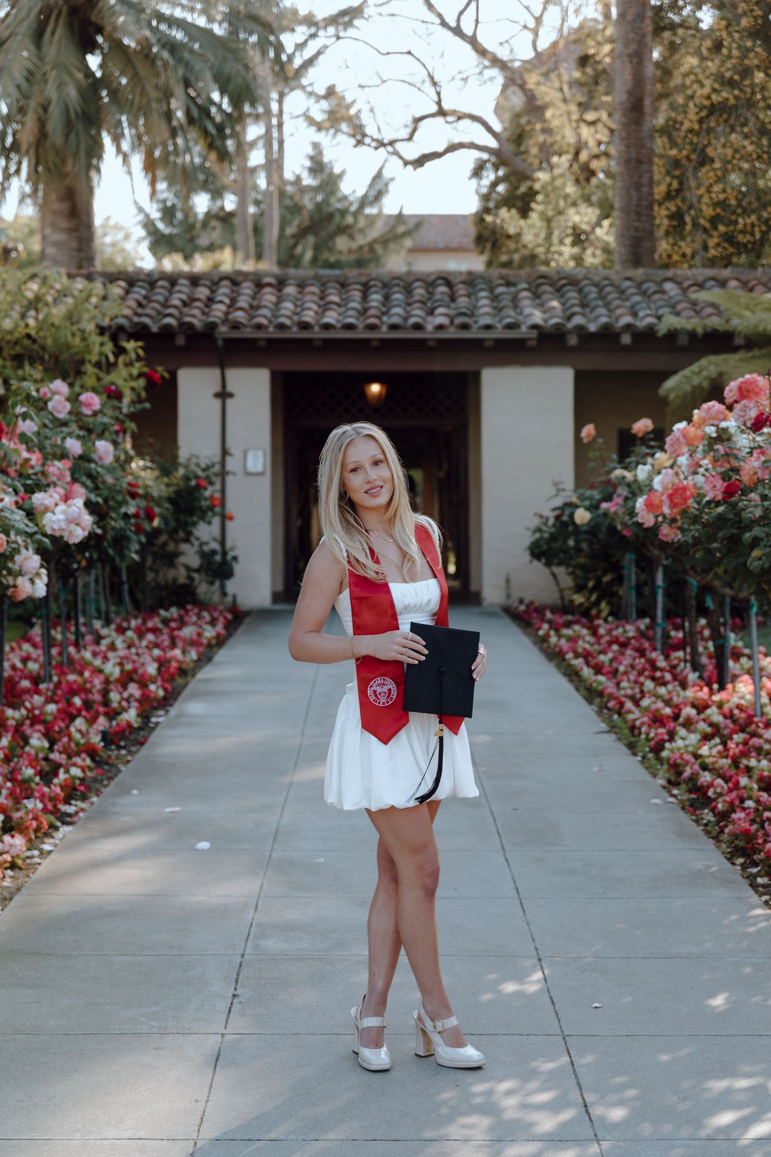 Santa Clara University graduate poses for her grad photoshoot by standing with rows of roses. Taken by Bay Area Graduation Photographer.