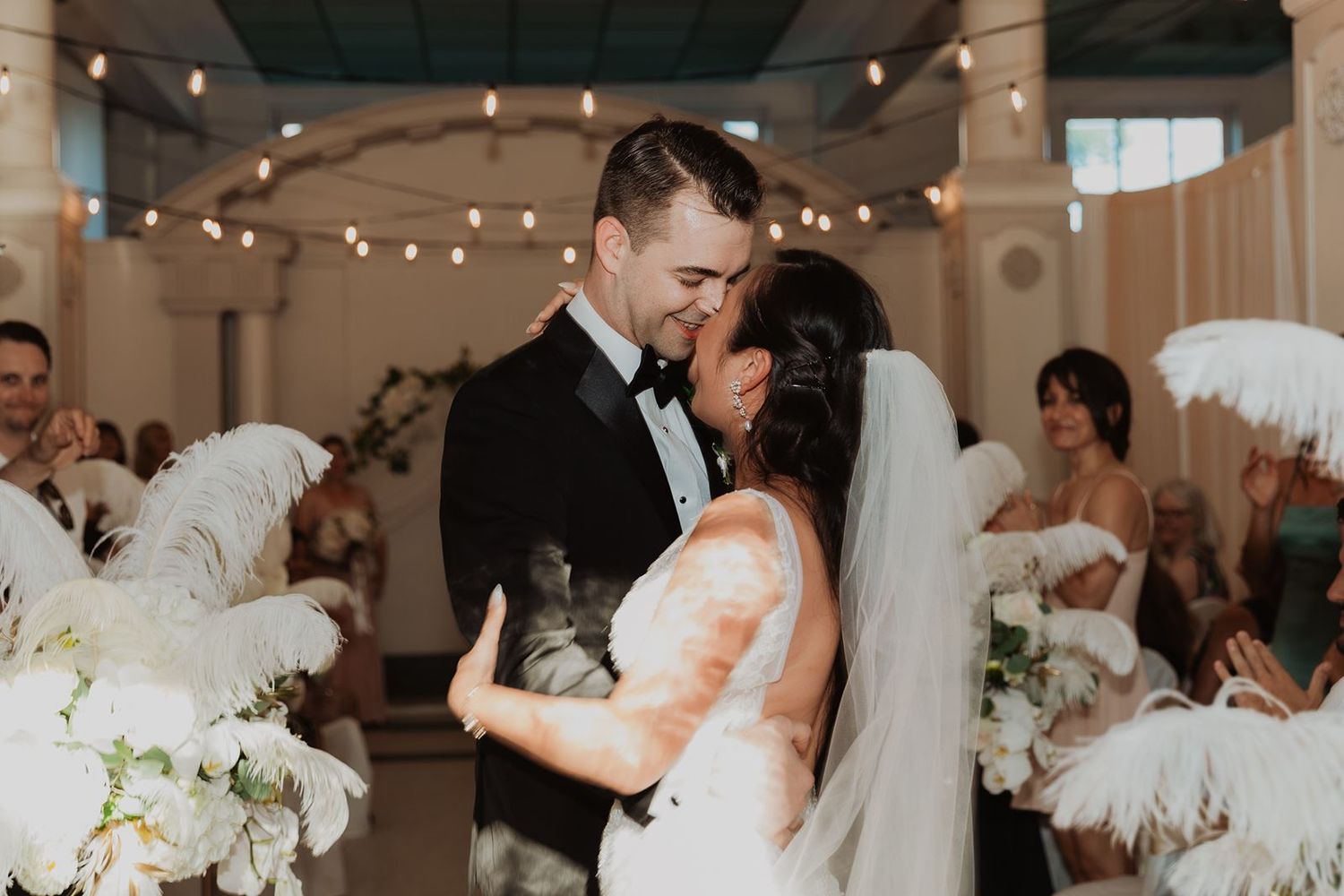 Bride and groom share first kiss at wedding ceremony surrounded by white feather fans and florals.