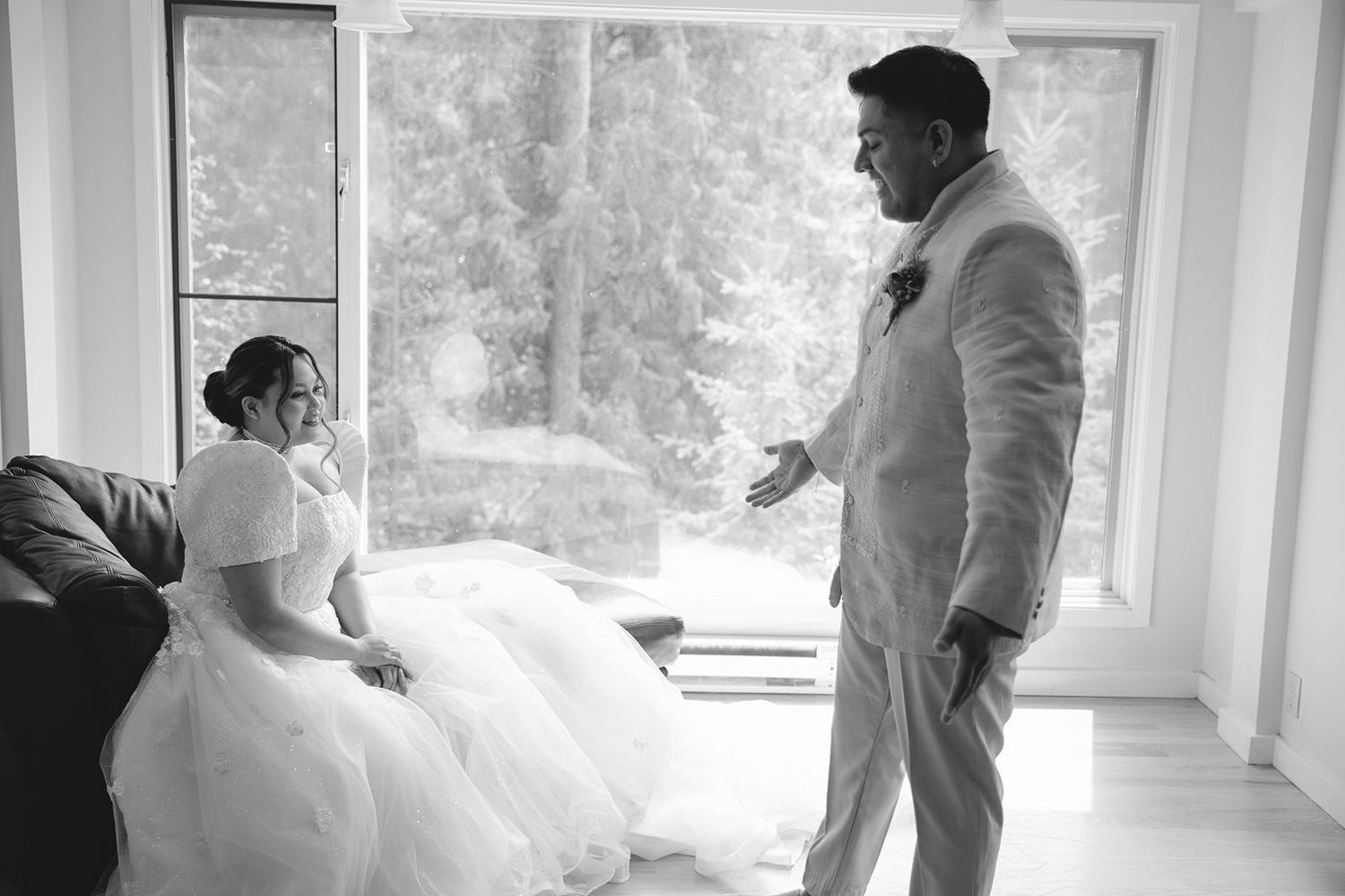 Black and white photo of groom in suit seeing bride in ballgown for the first time in a bright windowed room.