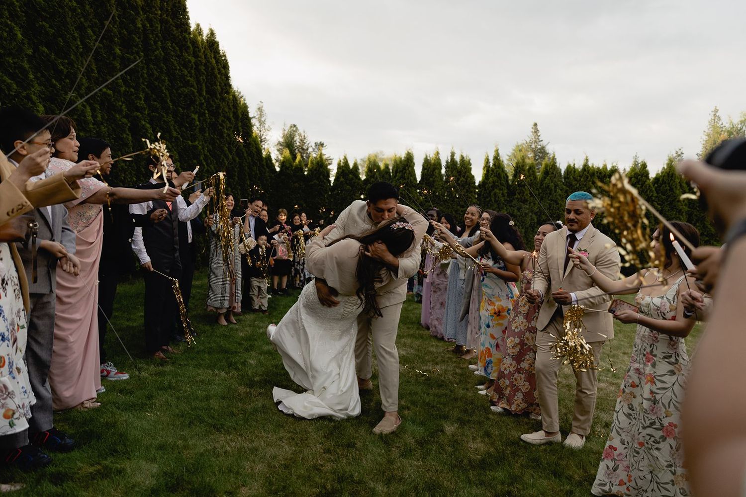 Groom dips bride for a kiss during a romantic sparkler exit surrounded by celebrating wedding guests outdoors.