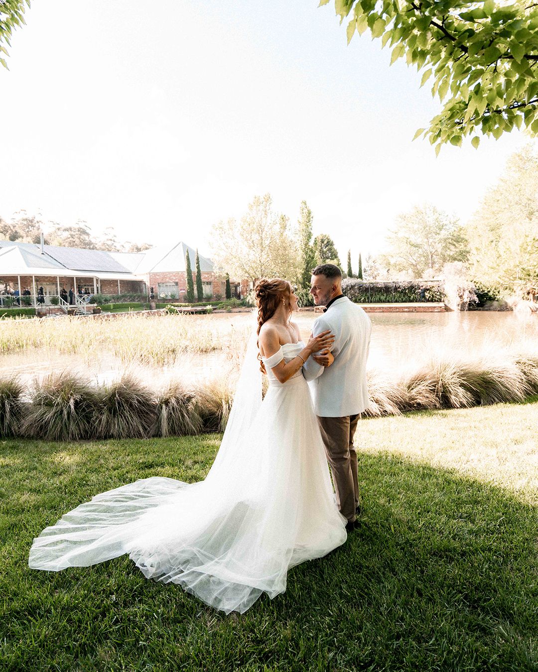 A couple in wedding attire dance together on a sunlit lawn with flowing white dress and veil creating elegant movement.
