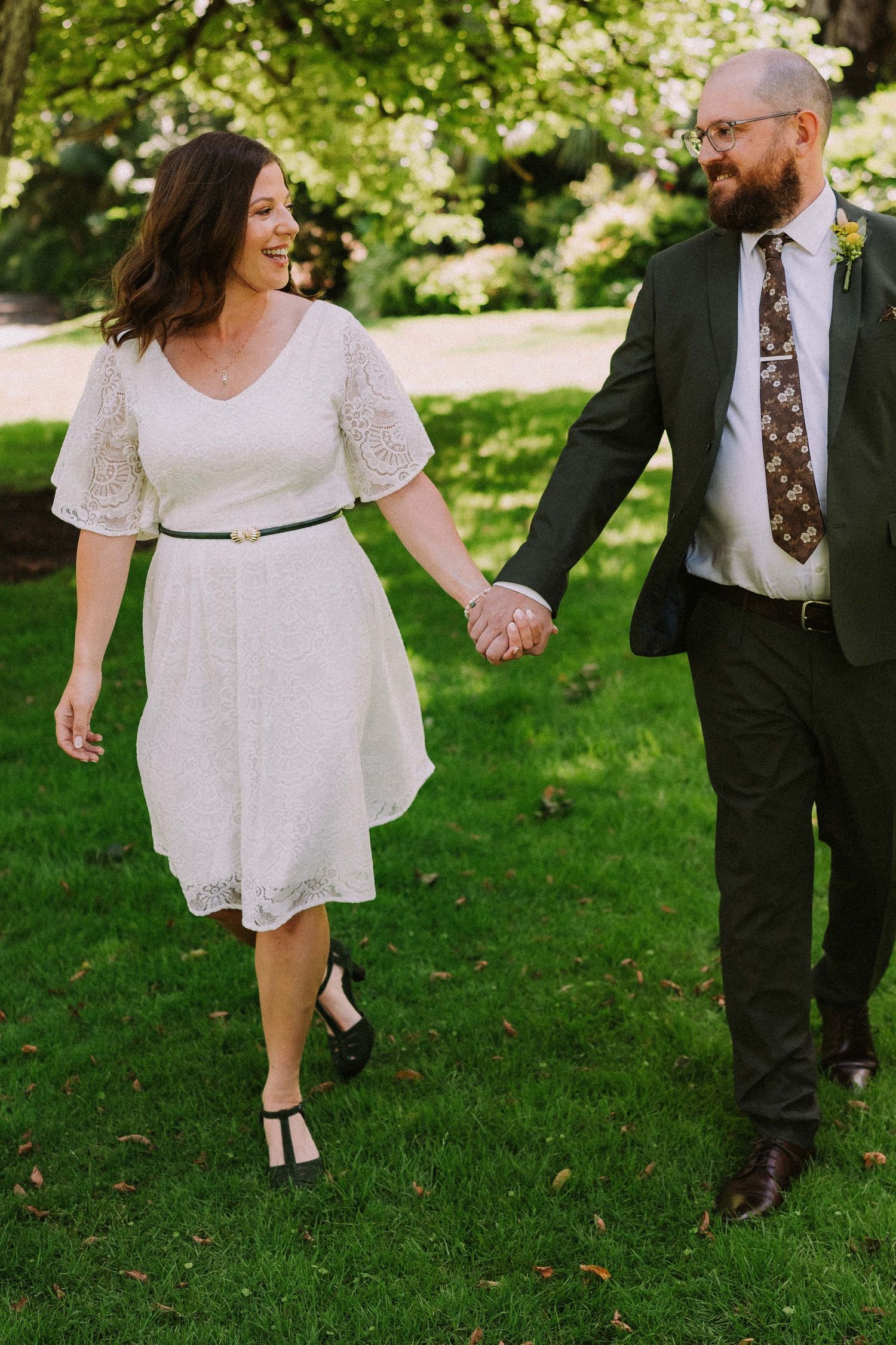 A couple in wedding attire playfully walks through a lush green garden setting on their special day.
