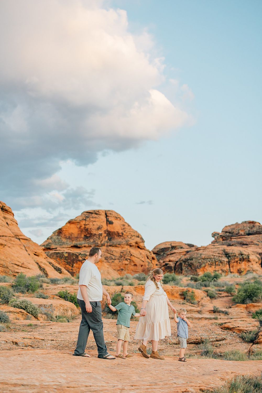 Family group walking together across scenic red rock desert formations under a blue sky with white clouds.