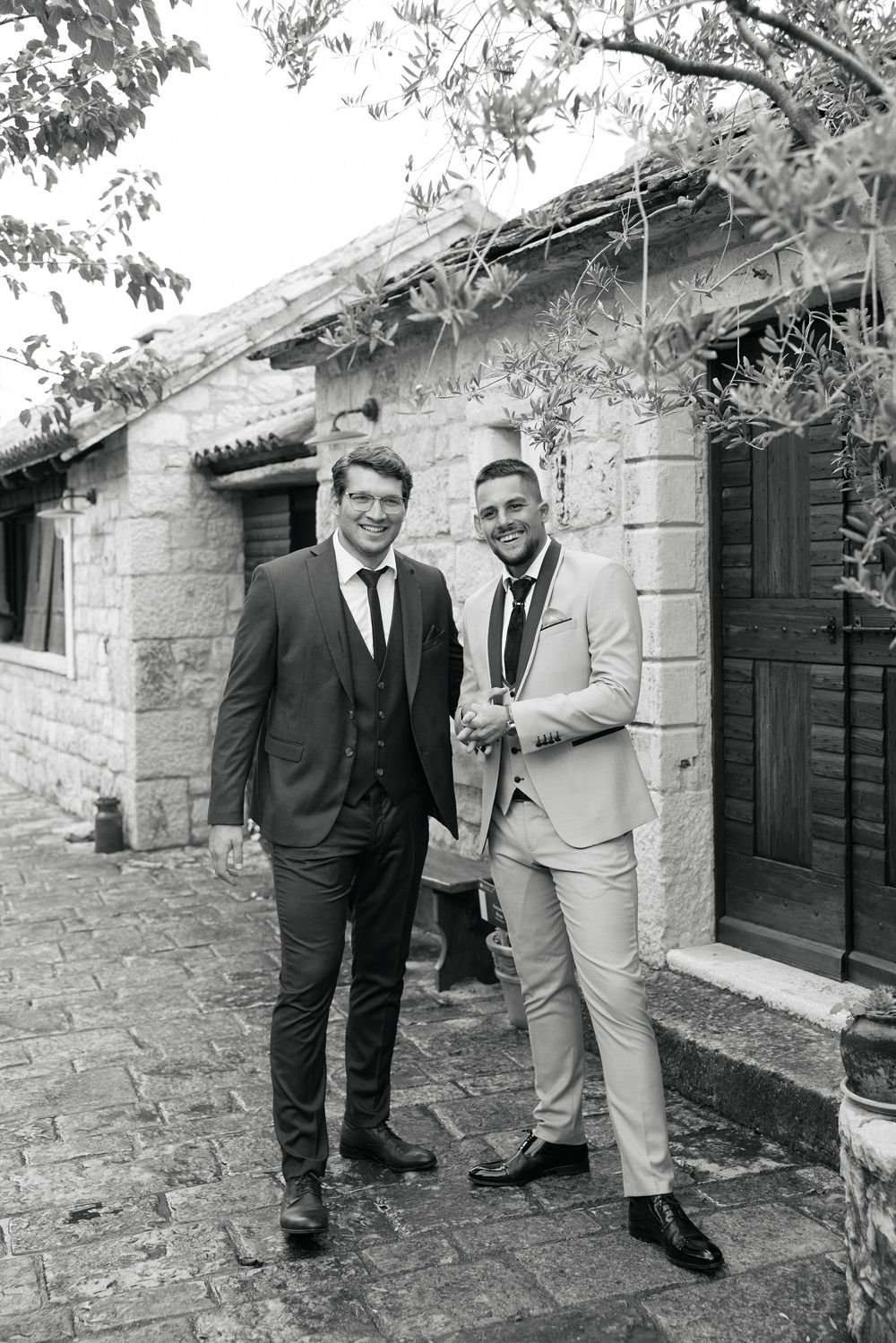 Two men in formal attire standing outside a stone building with black shutters.
