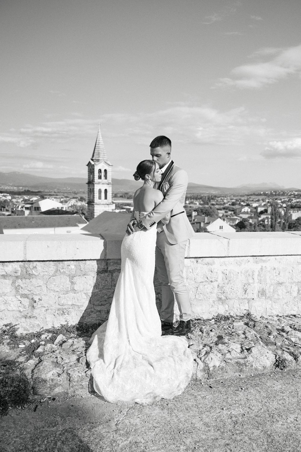 Black and white landscape photograph showing a scenic overlook with stone wall and church tower in the distance.
