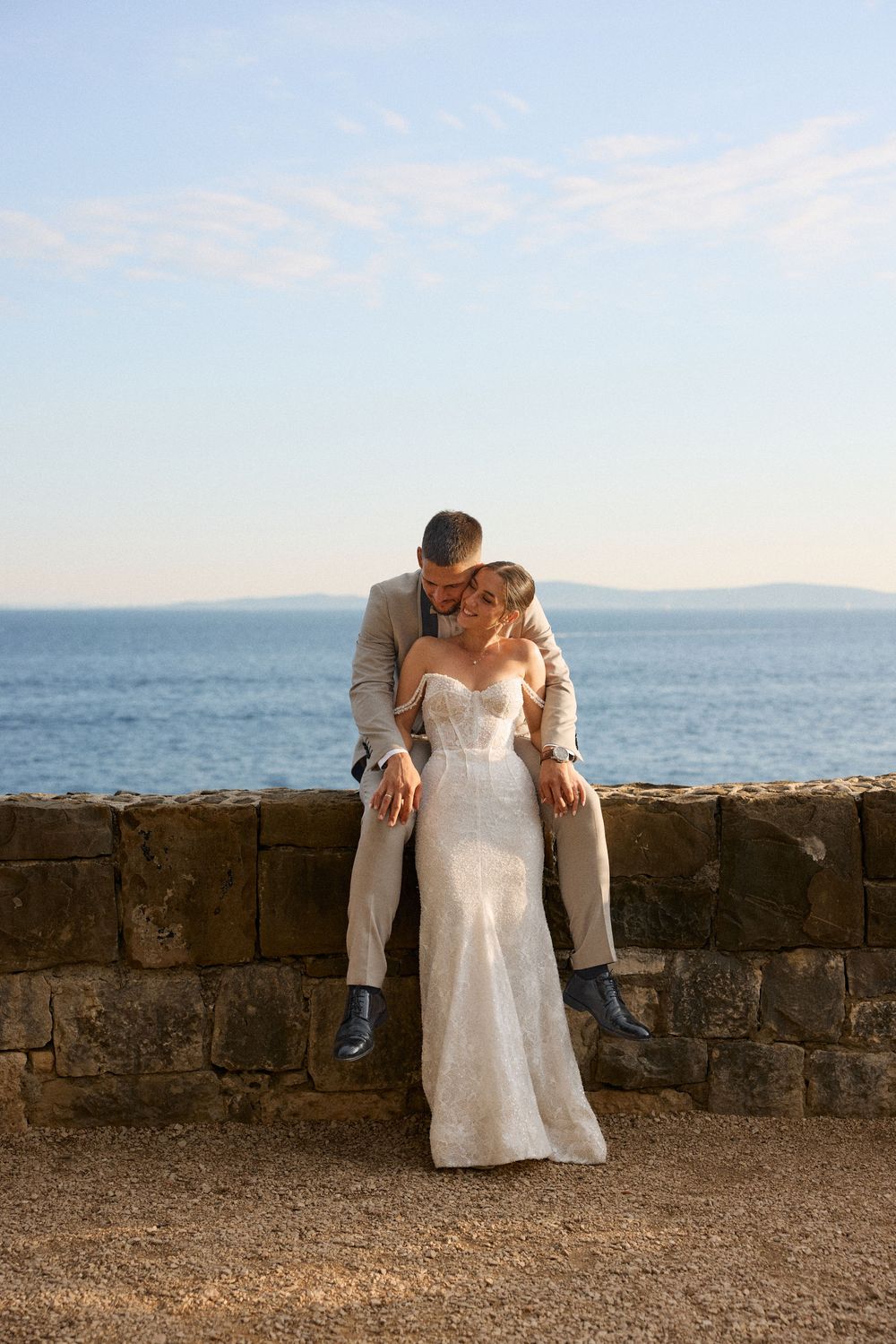 A romantic wedding couple sits on a stone wall overlooking the ocean at sunset.