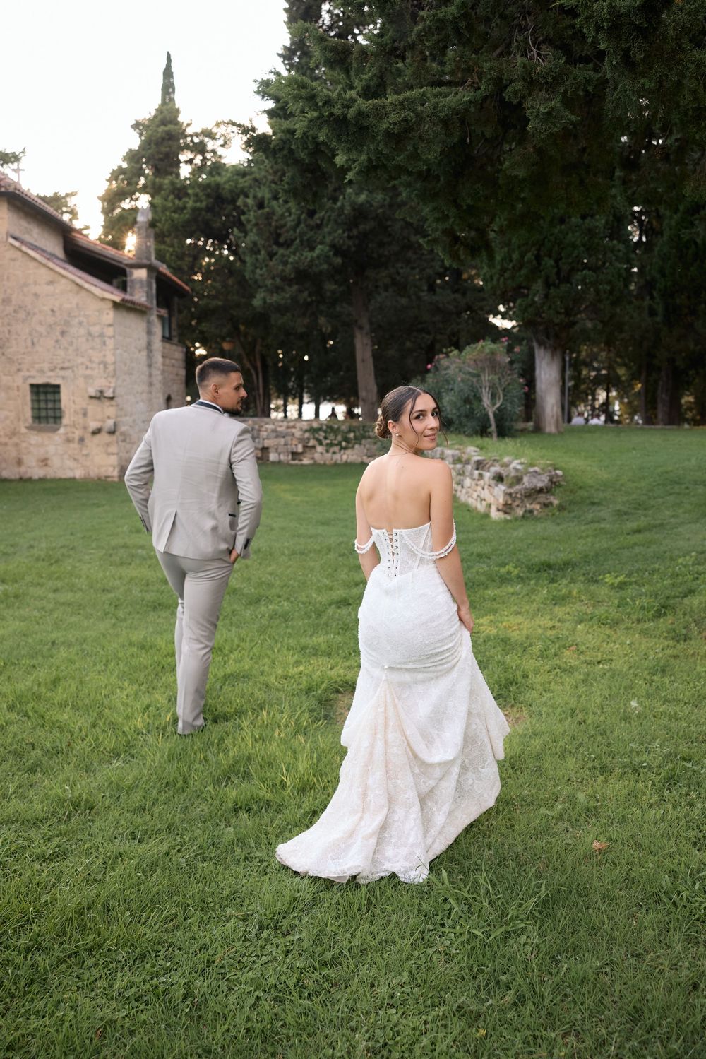 Wedding couple walking together on lush green grass near historic stone buildings.