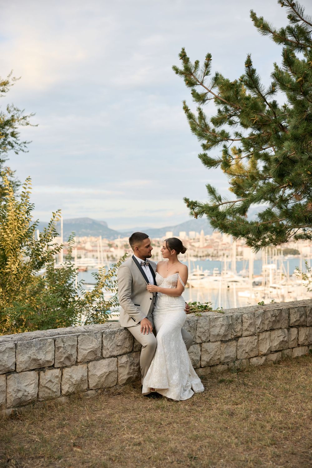 A romantic wedding couple shares an intimate moment overlooking a scenic cityscape from a stone wall viewpoint.