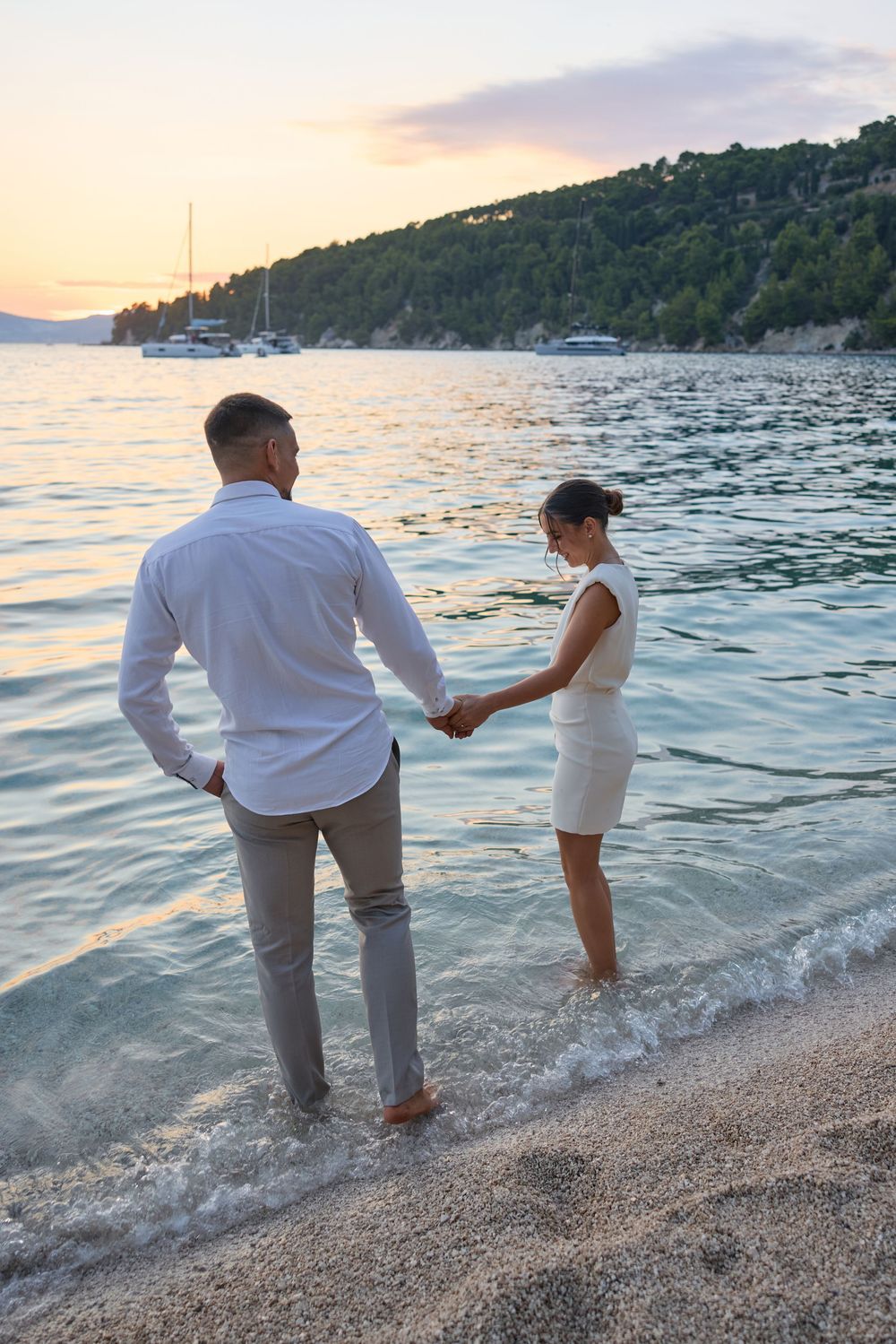 Romantic couple walking along beach shoreline at sunset holding hands.