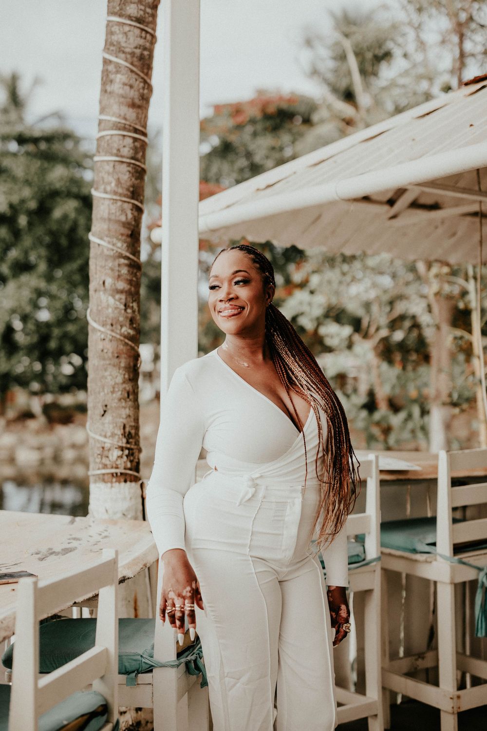 A joyful person in a white outfit poses outdoors against a backdrop of palm trees and patio umbrellas.