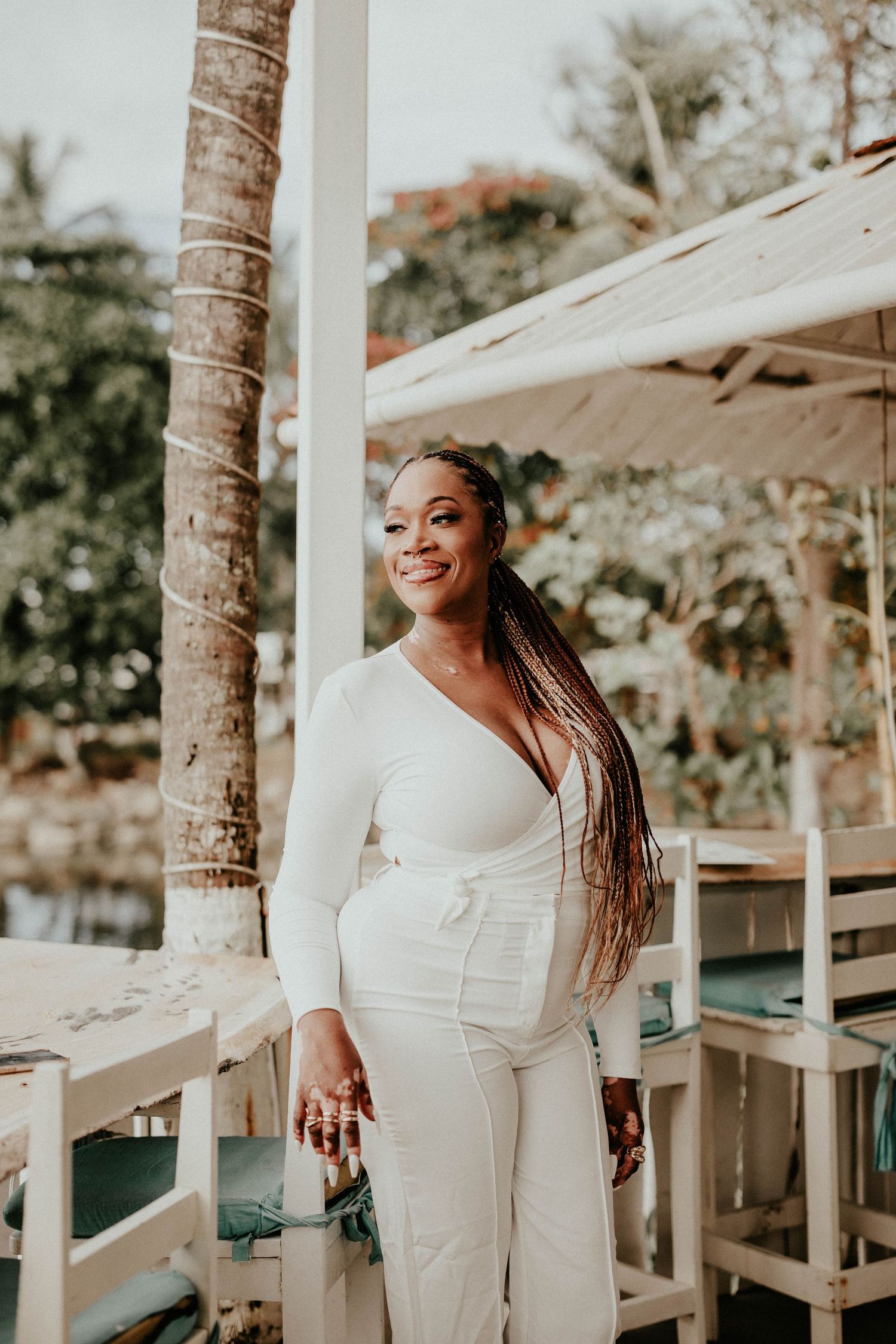 A joyful person in a white outfit poses outdoors against a backdrop of palm trees and patio umbrellas.
